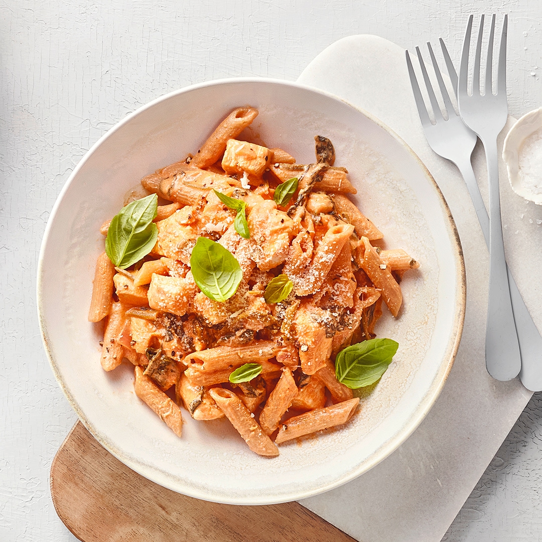 Schale mit Penne-Nudeln in cremiger Sauce, garniert mit Basilikum und geriebenem Käse; daneben Besteck und eine kleine Schale Parmesan.