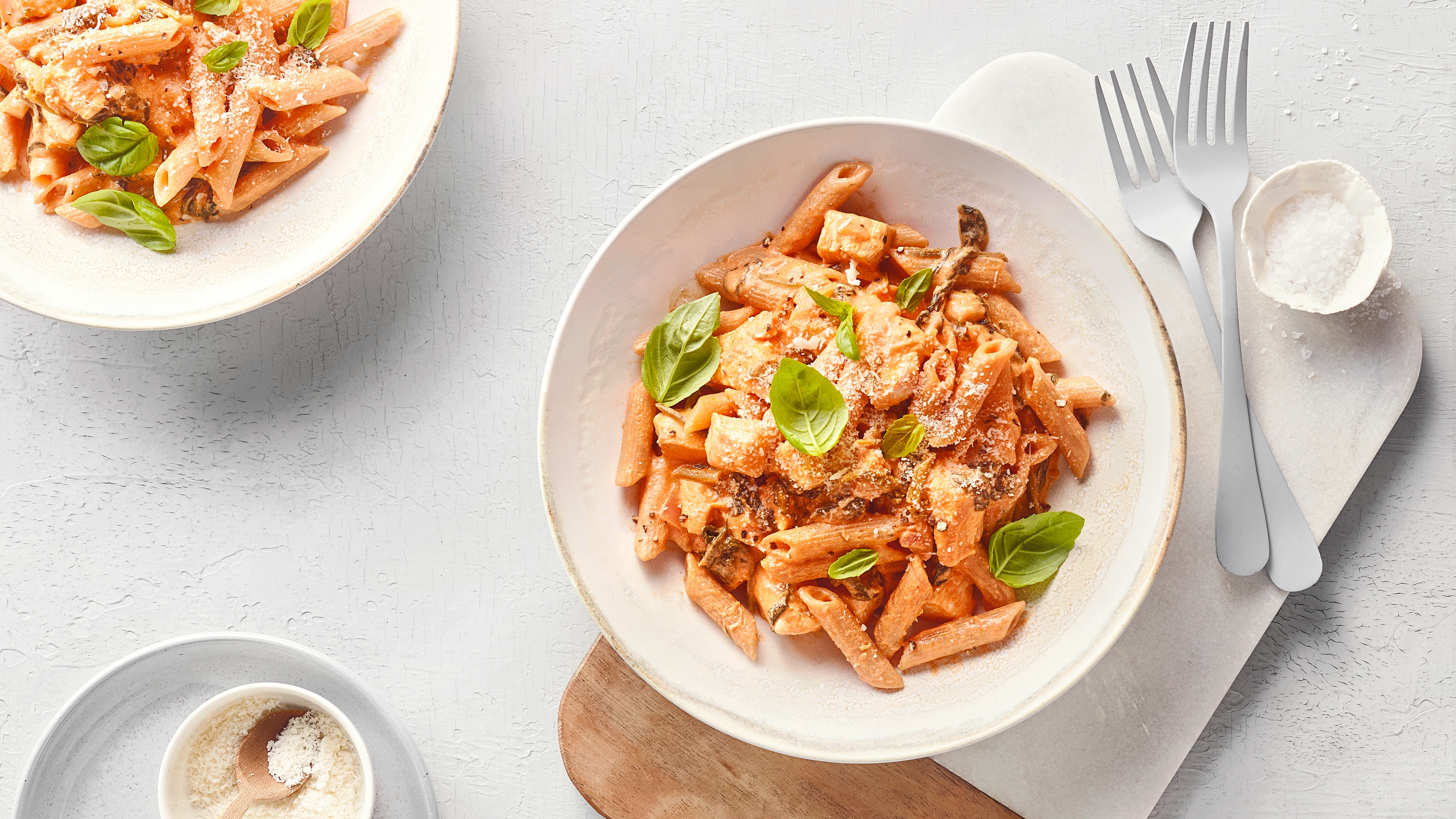 Schale mit Penne-Nudeln in cremiger Sauce, garniert mit Basilikum und geriebenem Käse; daneben Besteck und eine kleine Schale Parmesan.