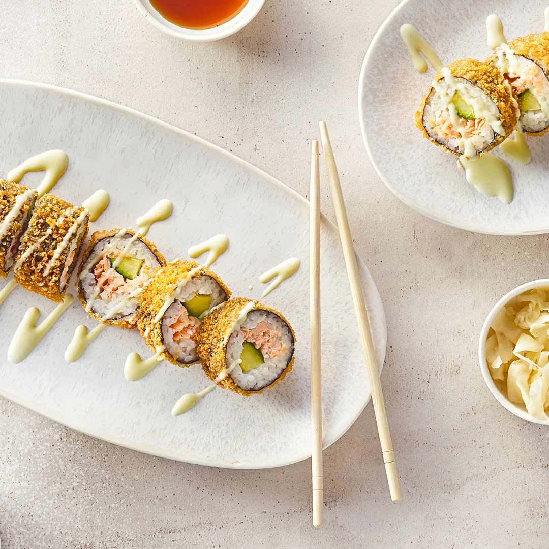 Sushi, umwickelt mit Noriblättern, paniert, gefüllt mit Reis, Gurke und Lachs, getoppt mit heller Soße, auf einem Teller