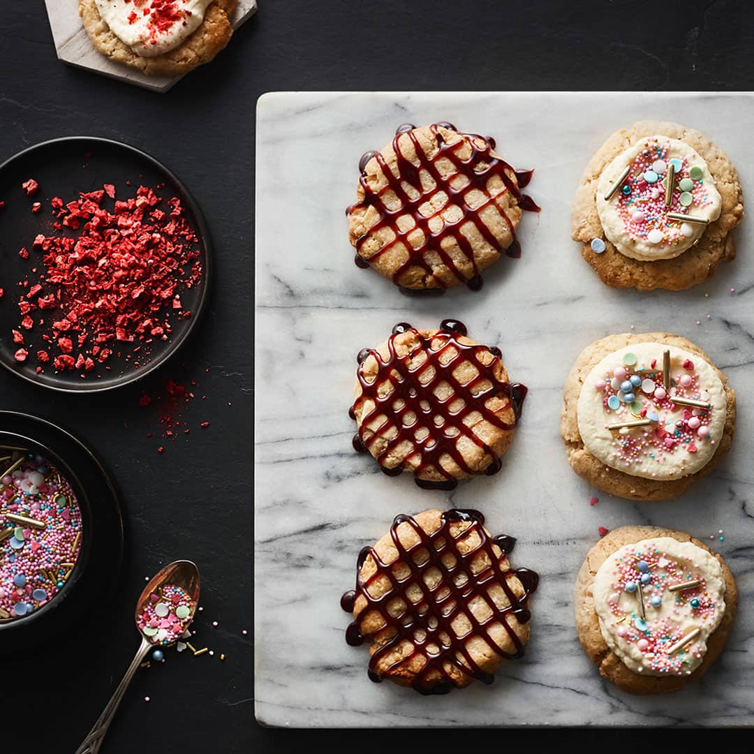 Große bunte Kekse, drei dekoriert mit dunklem Sirup, drei getoppt mit Frosting und Streuseln, auf einer hellen Marmorplatte.