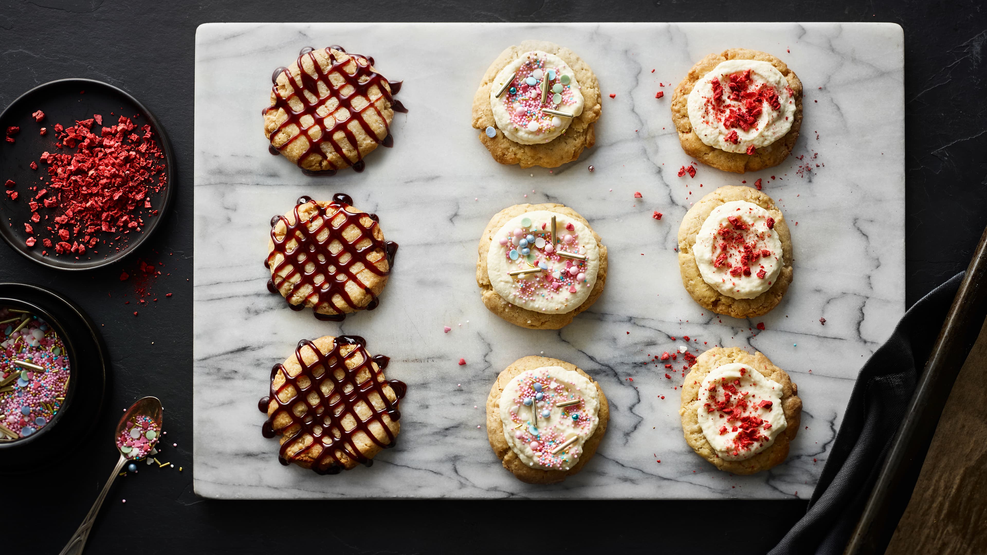 Große bunte Kekse, drei dekoriert mit dunklem Sirup, drei getoppt mit Frosting und Streuseln, drei mit Frosting und gefriergetrockneten Himbeeren, auf einer hellen Marmorplatte.