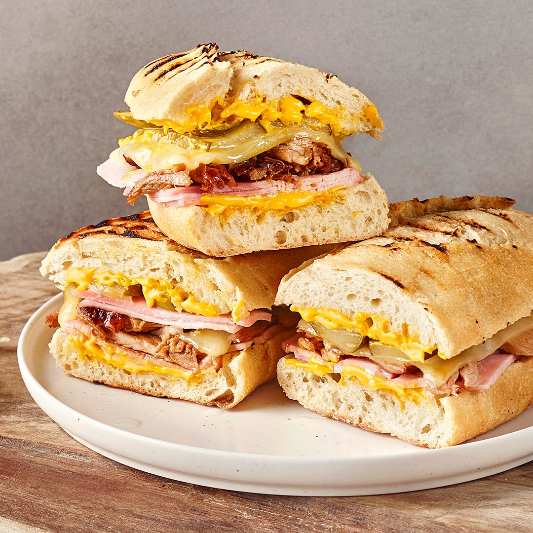 Ein Teller mit drei belegten Sandwichhälften, gefüllt mit Käse, Schinken, gebratenem Fleisch & eingelegten Gurkenscheiben.