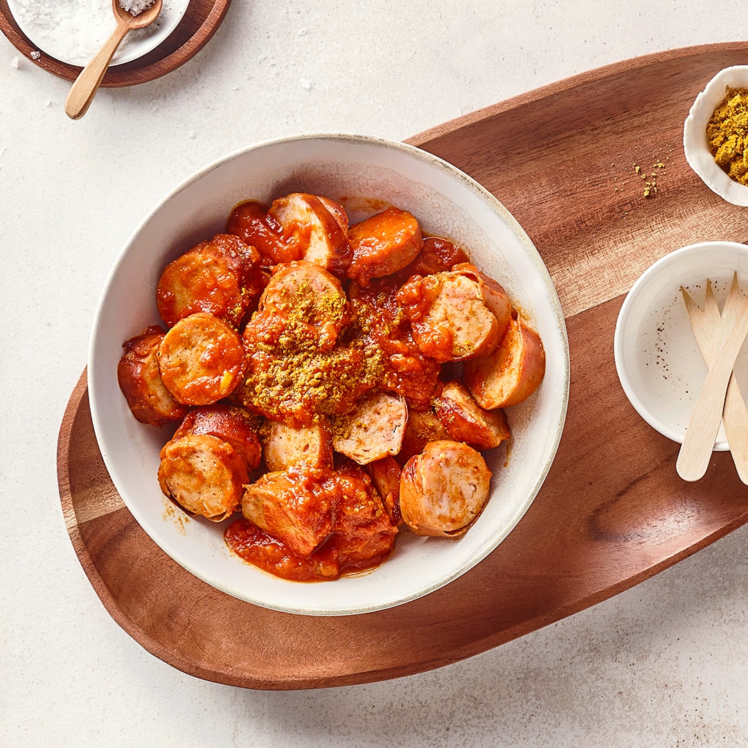 Eine Schale mit aufgeschnittenen Bratwürsten in roter Soße, bestreut mit gelbem Currypulver.