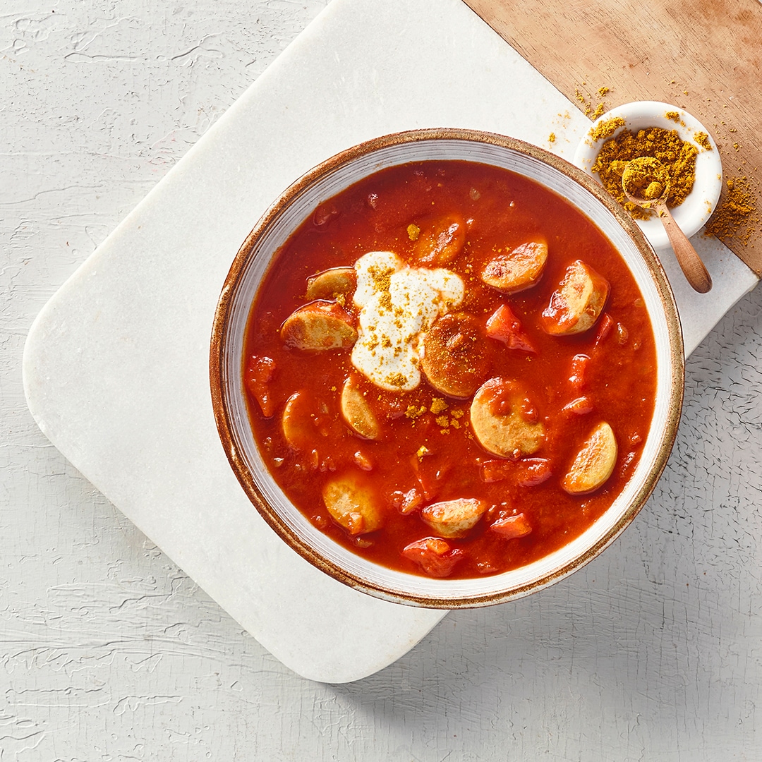 Eine Schale mit roter Soße, darin helle Wurstscheiben und rote Stücke, oben ein Klecks weißer Creme und gelbes Pulver.
