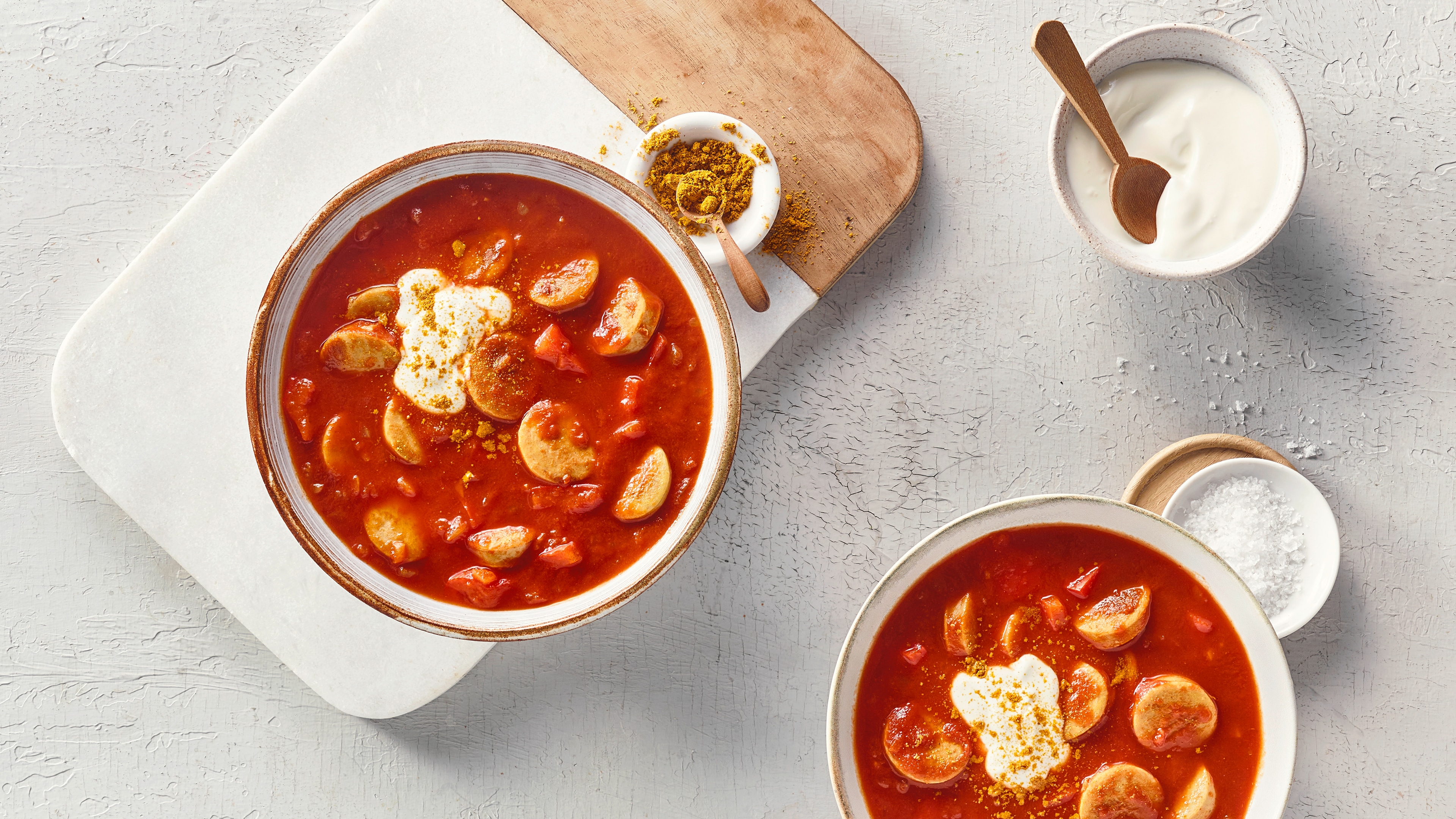 Eine Schale mit roter Soße, darin helle Wurstscheiben und rote Stücke, oben ein Klecks weißer Creme und gelbes Pulver.