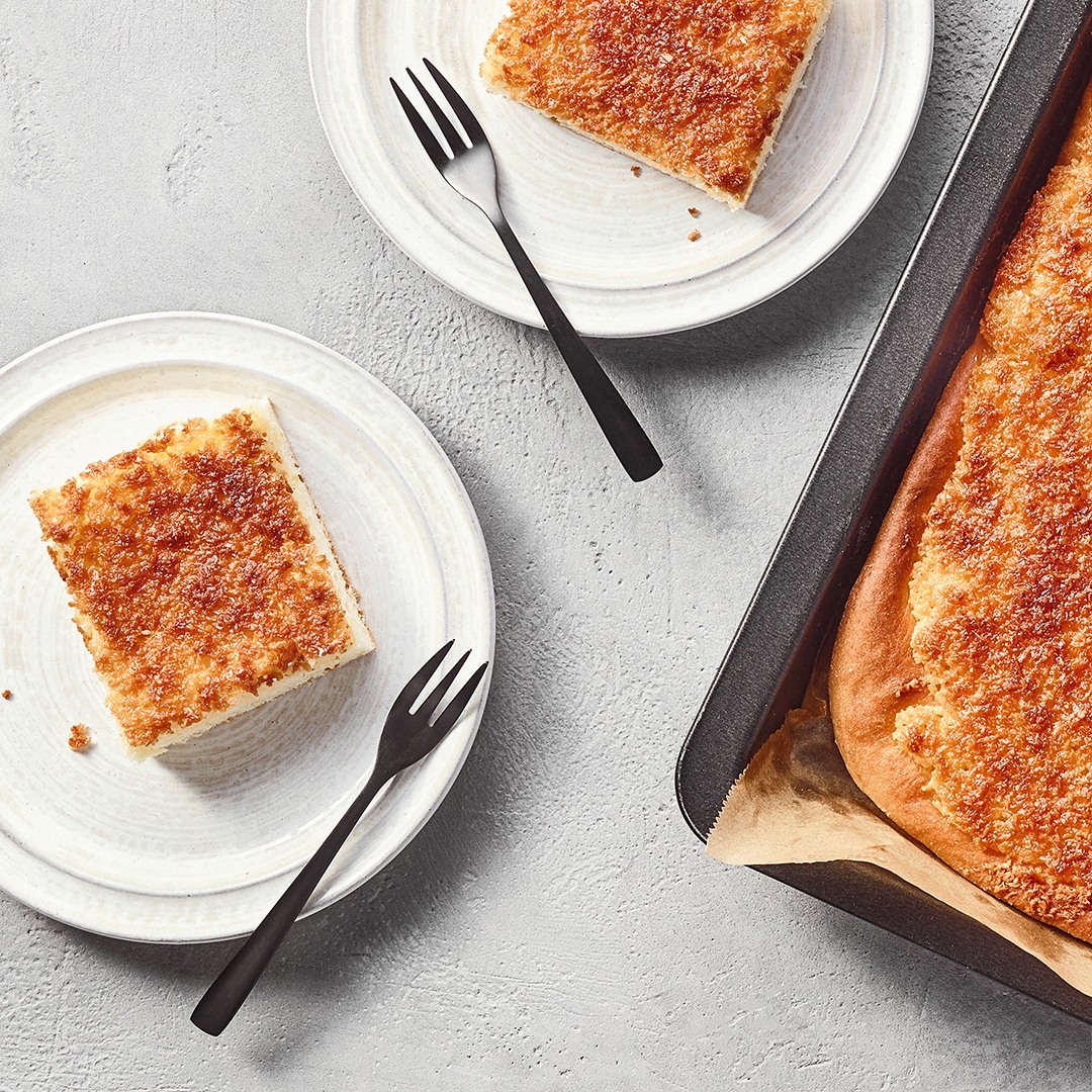 Ein Teller mit einem rechteckigen, hellen Kuchenstück mit goldbrauner Kruste, daneben ein Blech mit dem Kuchen.