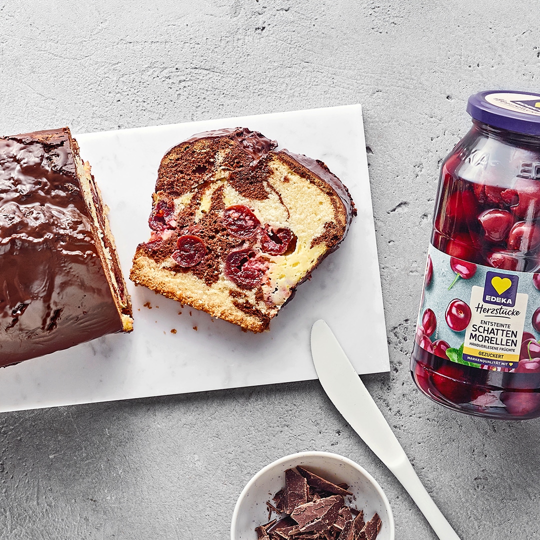 Kastenkuchen aus hellem und dunklem Rührteig und Sauerkirschen, überzogen mit Schokolade. Daneben EDEKA Herzstücke entsteinte Schattenmorellen.