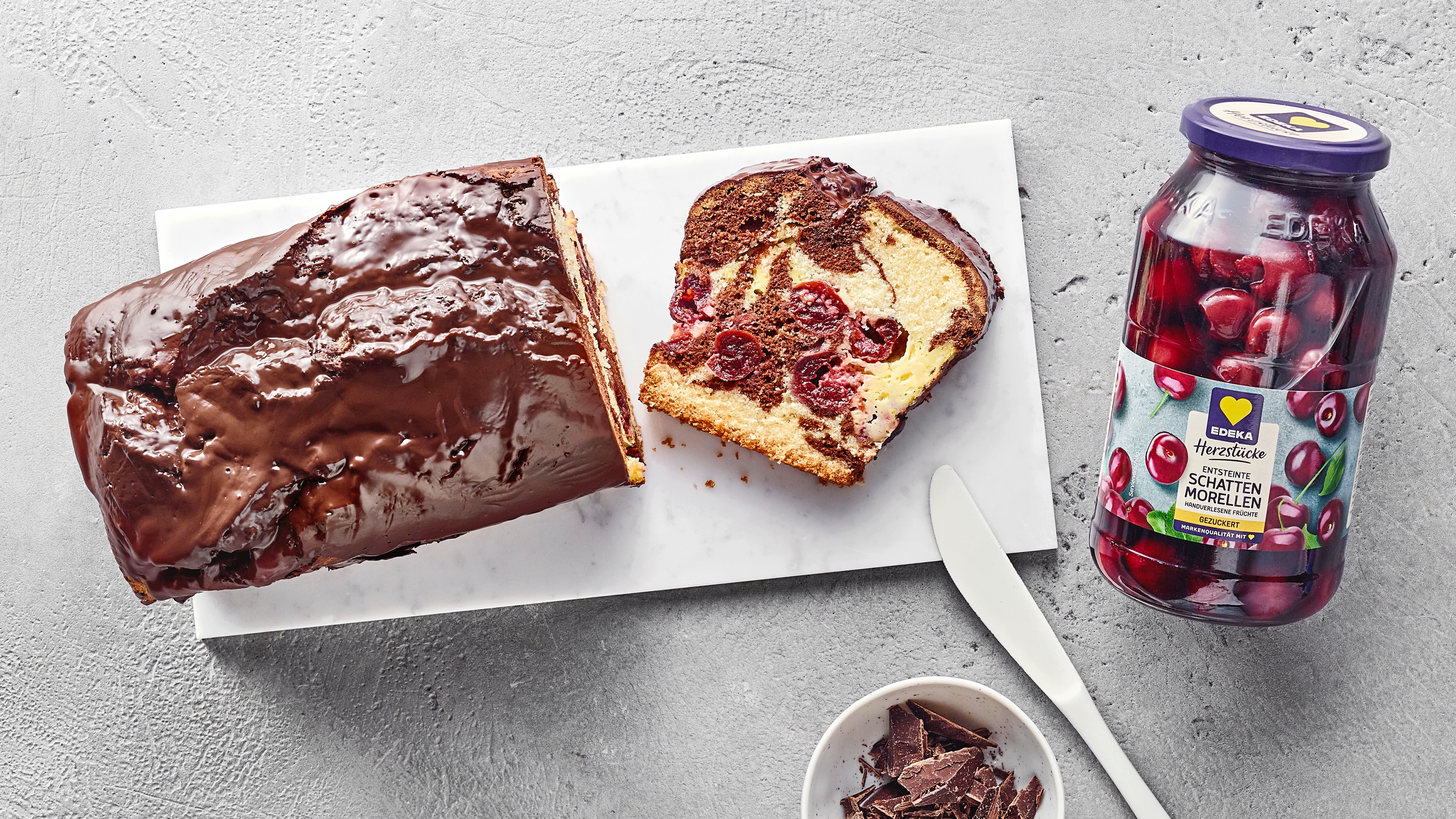 Kastenkuchen aus hellem und dunklem Rührteig und Sauerkirschen, überzogen mit Schokolade. Rechts daneben EDEKA Herzstücke entsteinte Schattenmorellen.