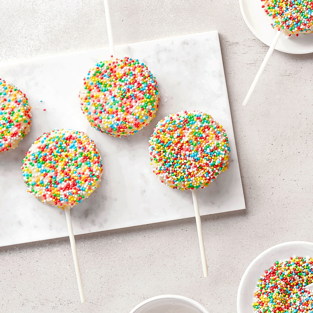Vier bunte Cake Pops mit Zuckerstreuseln auf einer weißen Marmorplatte, umgeben von Schalen mit bunten Streuseln.