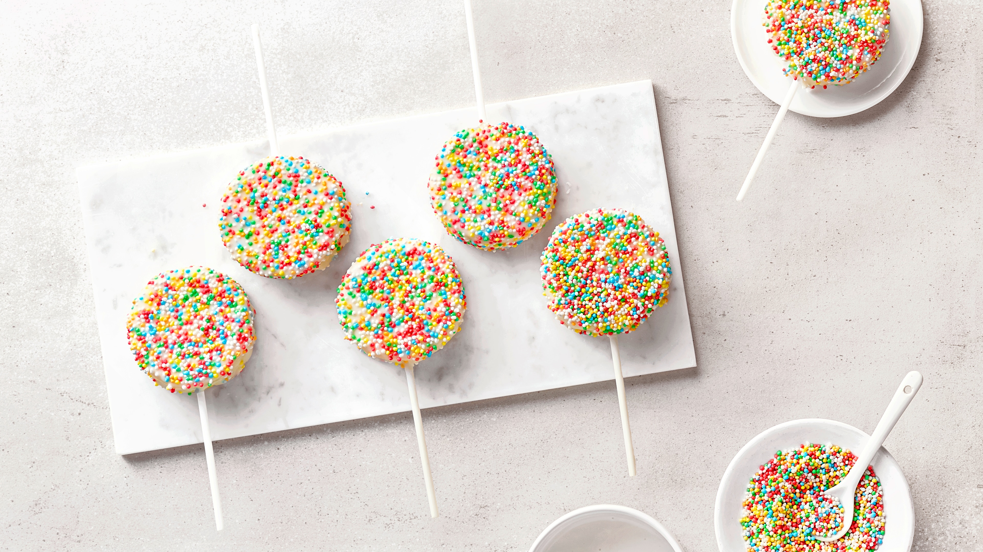 Vier bunte Cake Pops mit Zuckerstreuseln auf einer weißen Marmorplatte, umgeben von Schalen mit bunten Streuseln.
