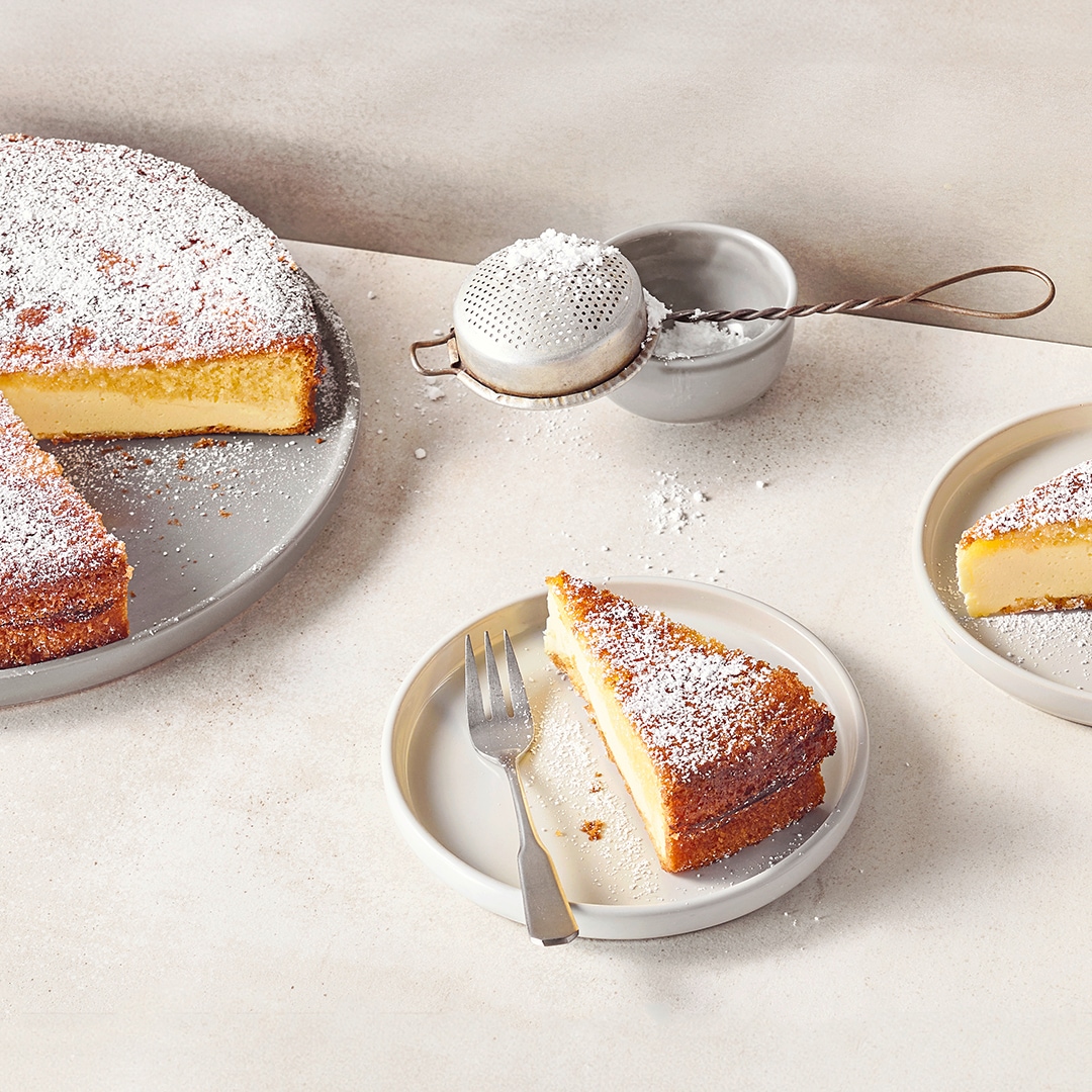 Teller mit Kuchenstücken aus einem hellen, cremigen Boden und einem fluffigen Teigdeckel, bestreut mit feinem Puderzucker