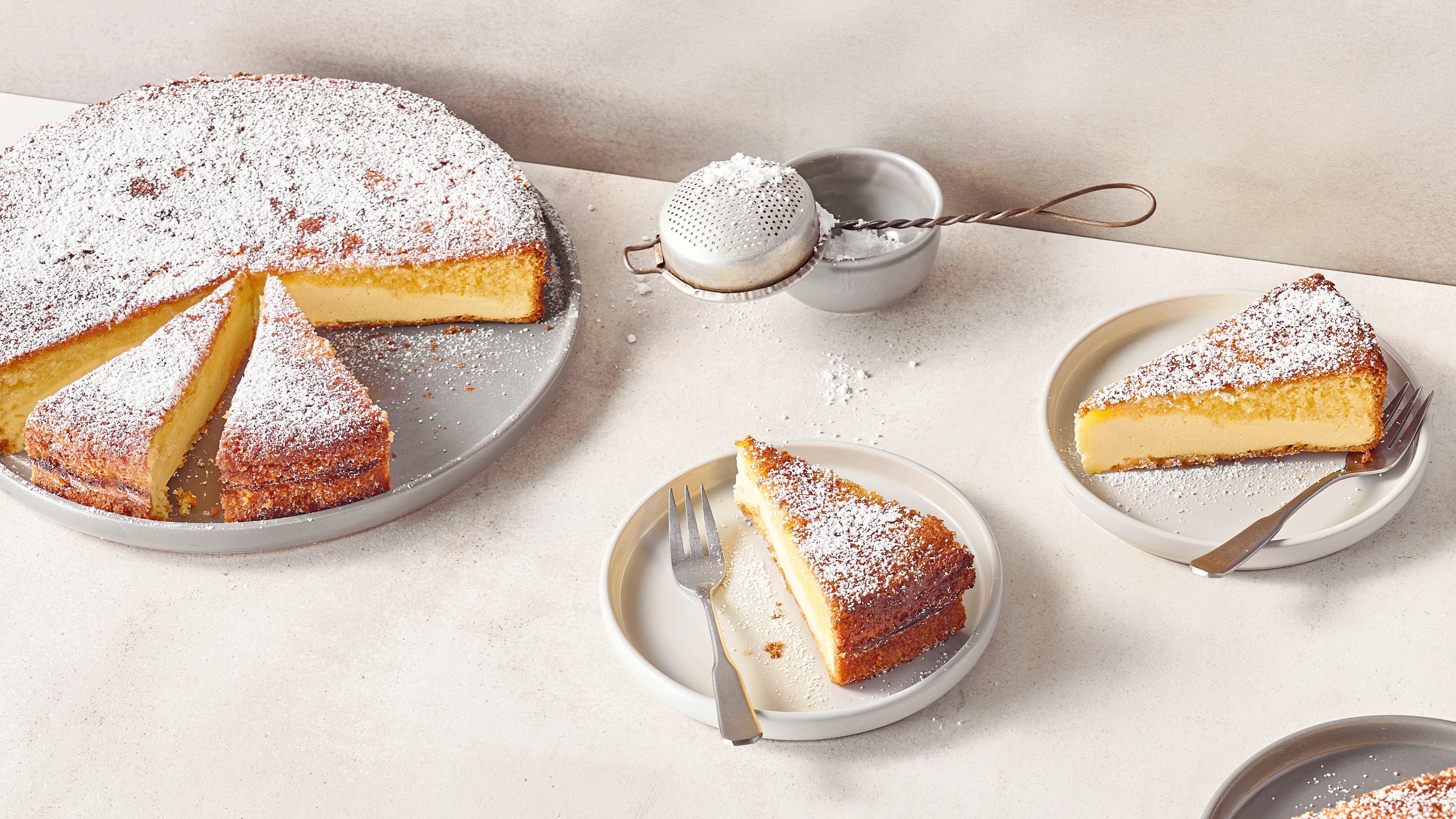 Rezept für Dreh-dich-um-Kuchen Teller mit Kuchenstücken aus einem hellen, cremigen Boden und einem fluffigen Teigdeckel, bestreut mit feinem Puderzucker