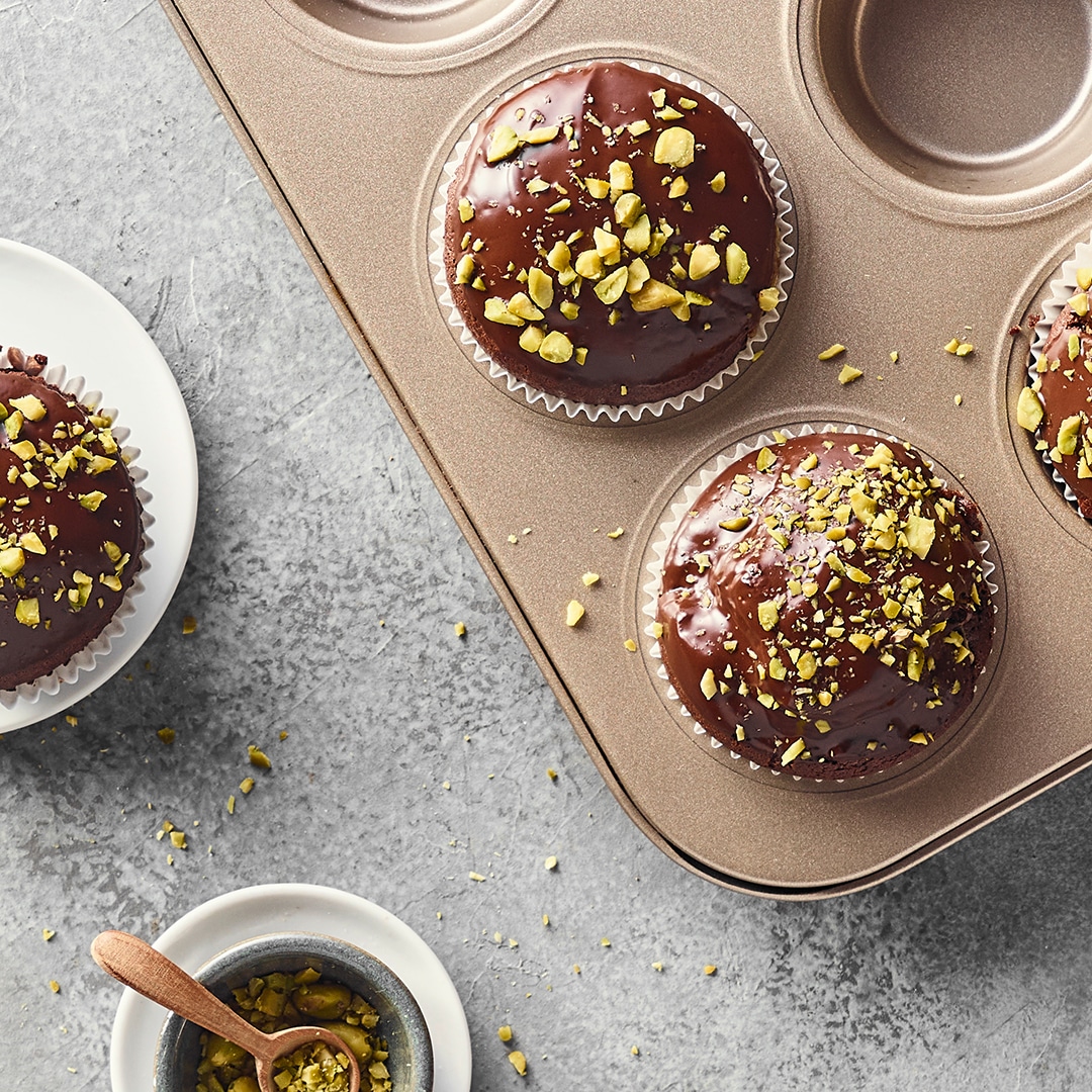 Ein Blech mit Schokoladenmuffins, bedeckt mit dunkler Glasur und garniert mit gehackten Pistazienstücken