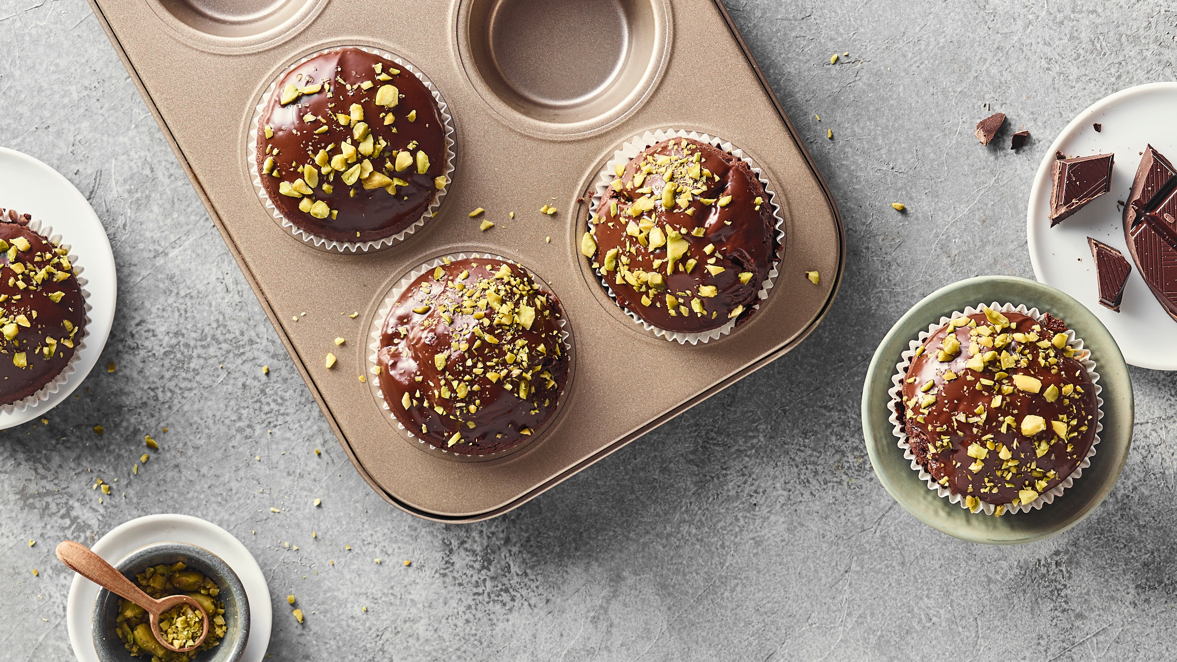 Ein Blech mit Schokoladenmuffins, bedeckt mit dunkler Glasur und garniert mit gehackten Pistazienstücken