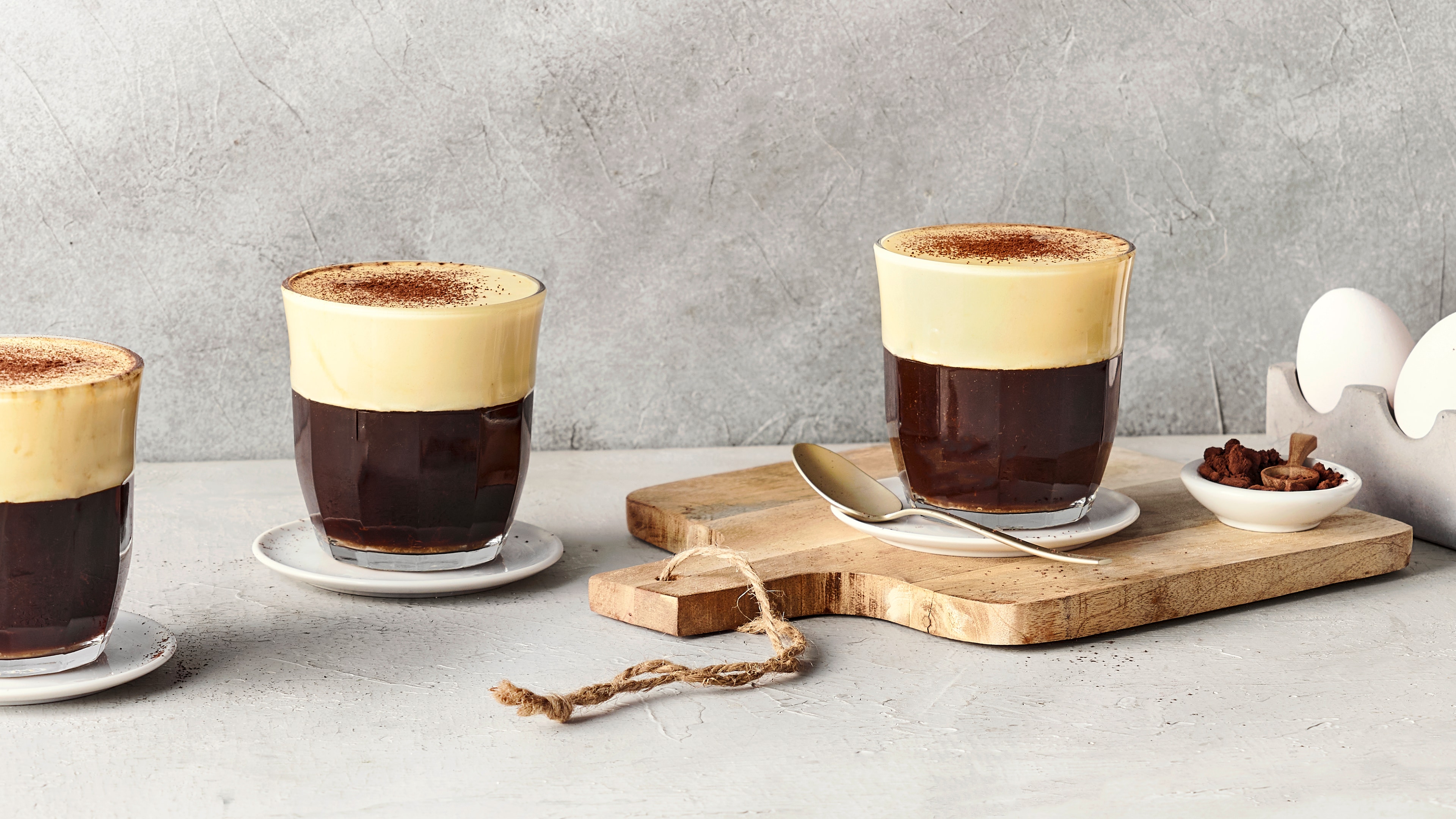 Ein Glas mit dunklem Kaffee, bedeckt mit heller Schaumschicht und bestreut mit feinem braunem Pulver, daneben ein Löffel