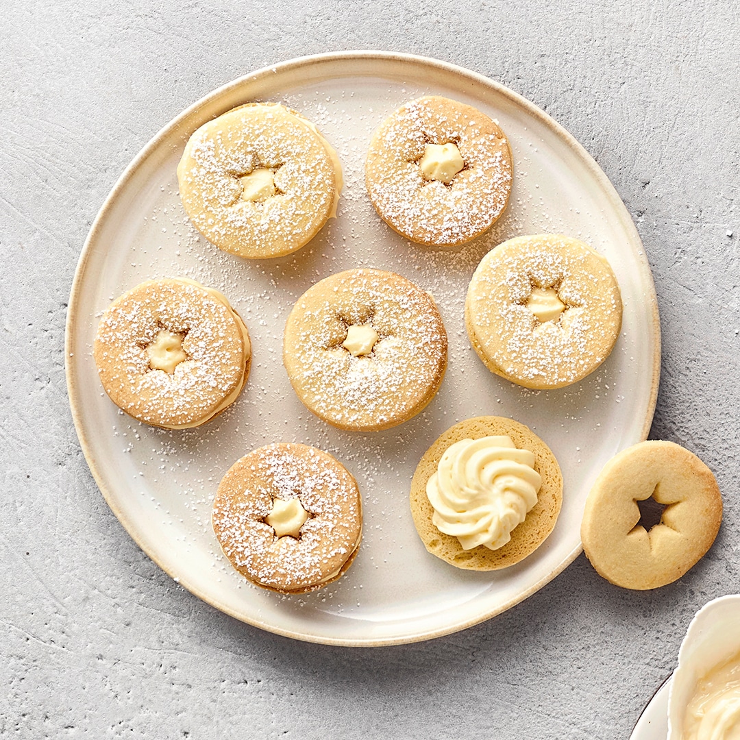 Runder, beigefarbener Teller mit sieben runden Plätzchen, jeweils mit sternförmigem Ausstecher und einer Eierlikör‑Cremefüllung. Mit Puderzucker bestäubt.