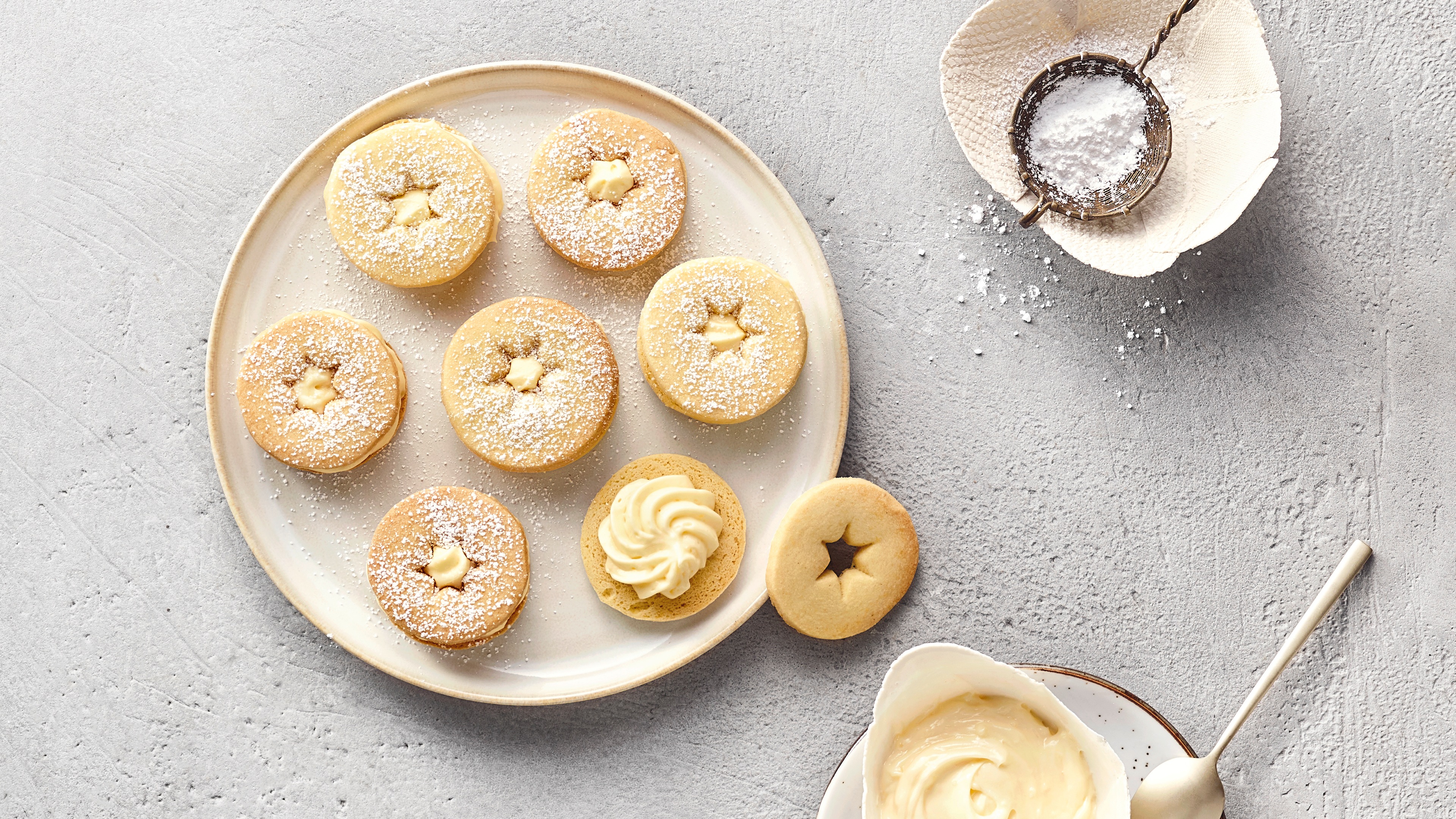 Runder, beigefarbener Teller mit sieben runden Plätzchen, jeweils mit sternförmigem Ausstecher und einer Eierlikör‑Cremefüllung. Mit Puderzucker bestäubt. Rechts daneben liegen kleine Schüsseln mit zusätzlichem Puderzucker und etwas Creme.