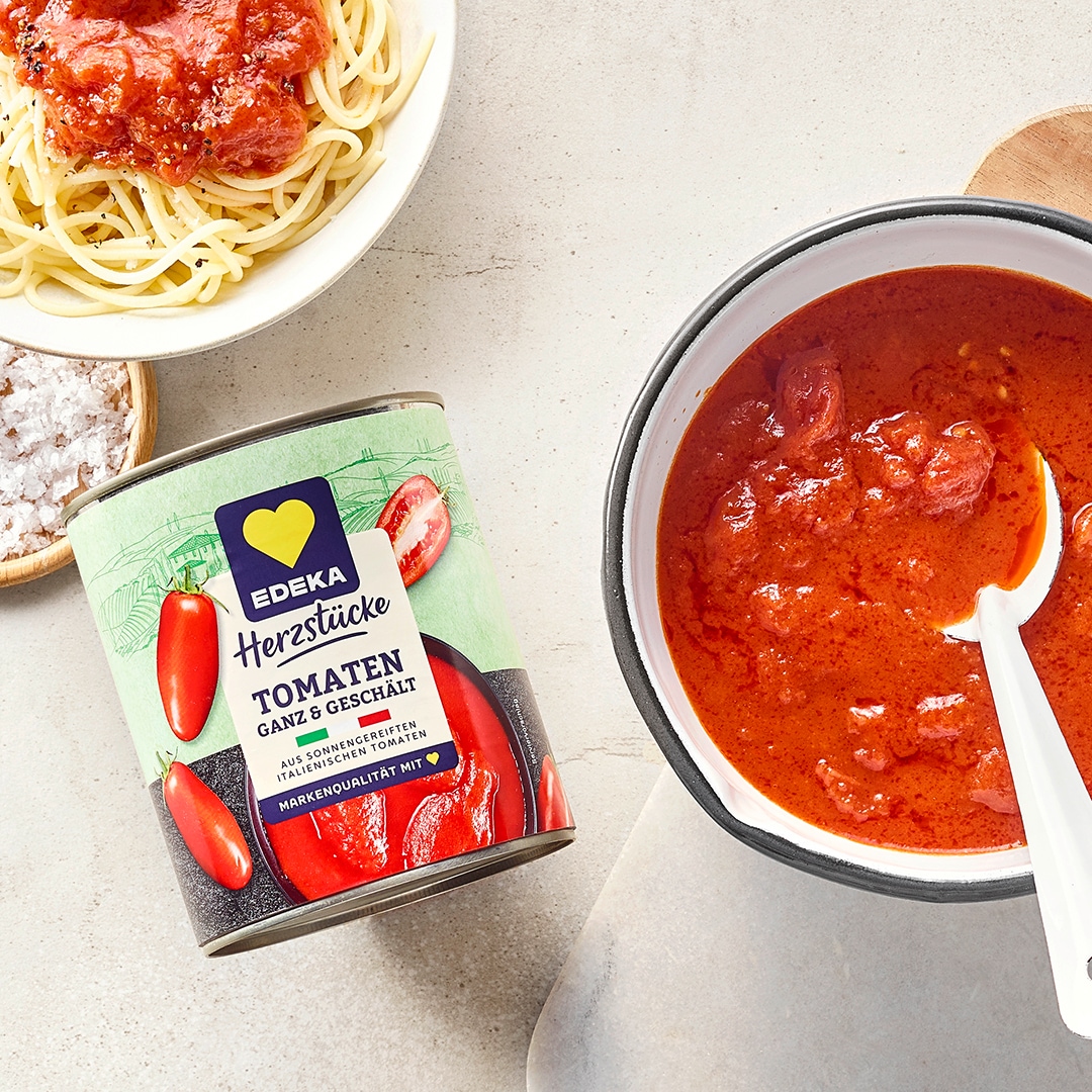 Schüssel mit roter, stückiger Tomatensauce und Löffel, daneben Spaghetti, Gewürze und eine kleine Tomatenschüssel. Links daneben EDEKA Herzstücke Tomaten ganz & geschält.
