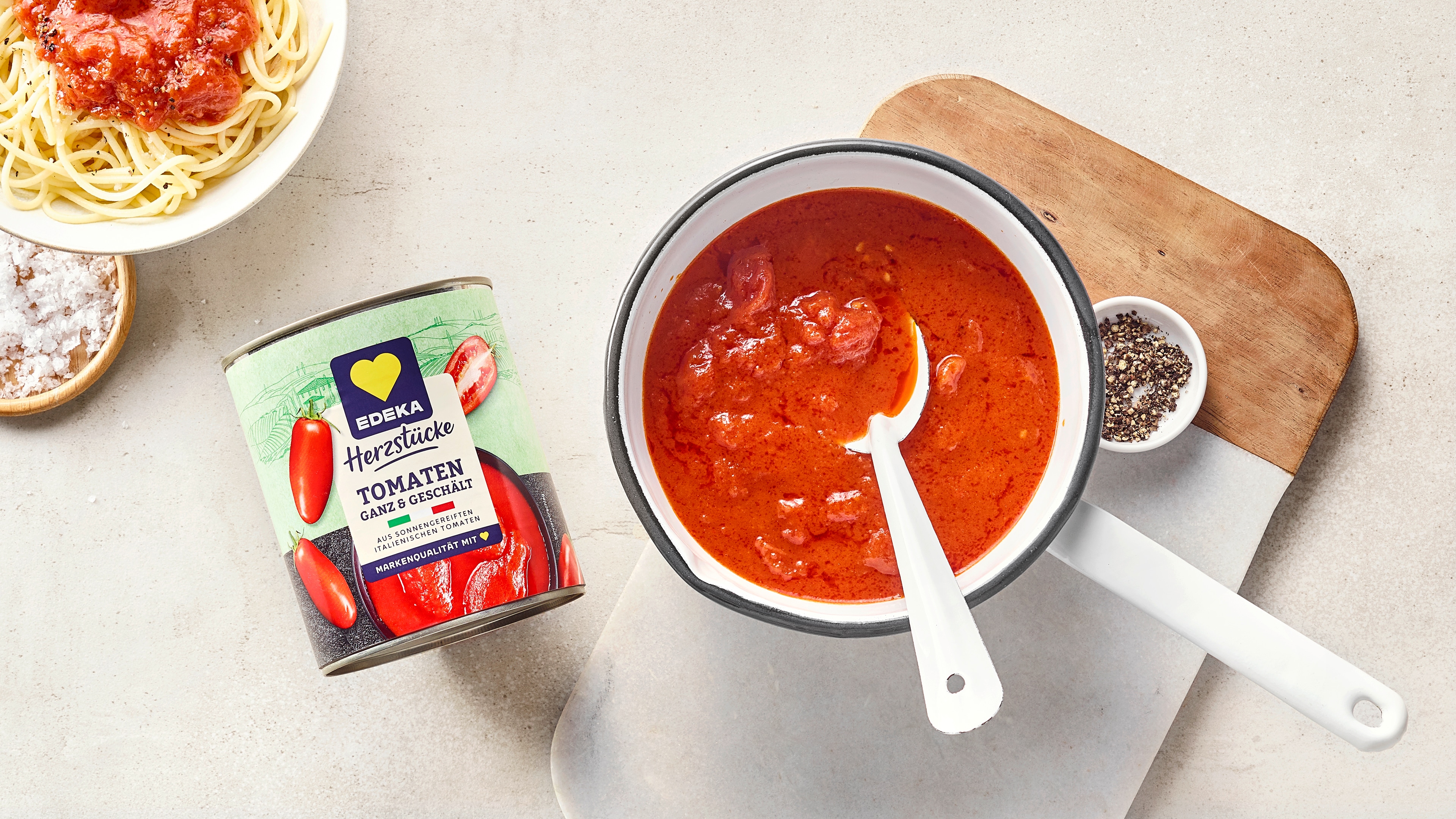 Schüssel mit roter, stückiger Tomatensauce und Löffel, daneben Spaghetti, Gewürze und eine kleine Tomatenschüssel. Links daneben EDEKA Herzstücke Tomaten ganz & geschält.