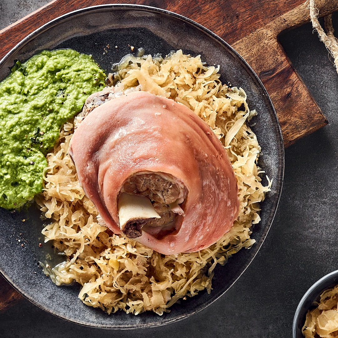 Ein Teller mit einem hellen, runden Stück Fleisch, gebettet auf Sauerkraut, dazu cremiges, kräftig grünes Püree