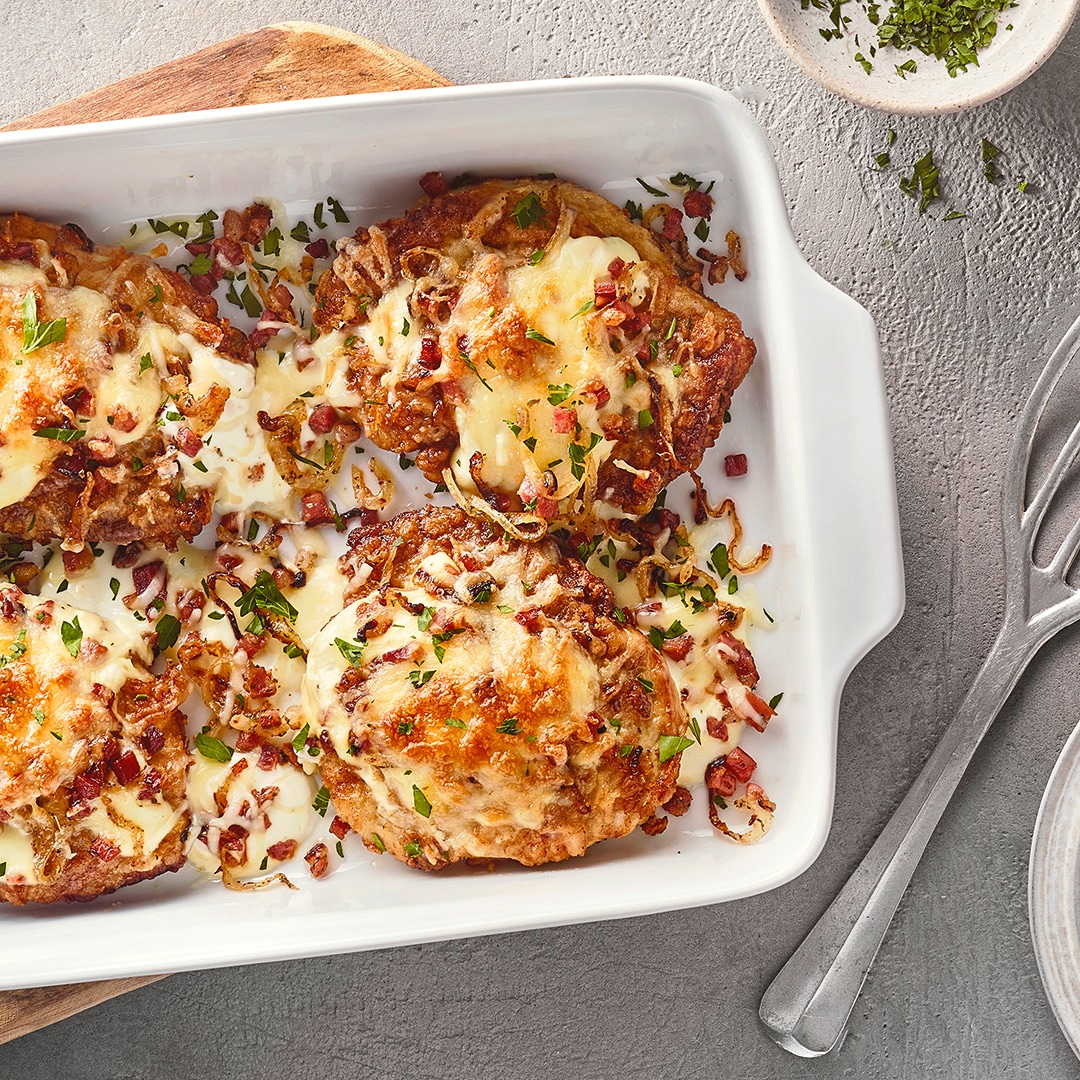 Eine Auflaufform mit vier gebräunten Fleischstücken, überbacken mit Käse, kleinen roten Würfeln & grünen Kräutern.