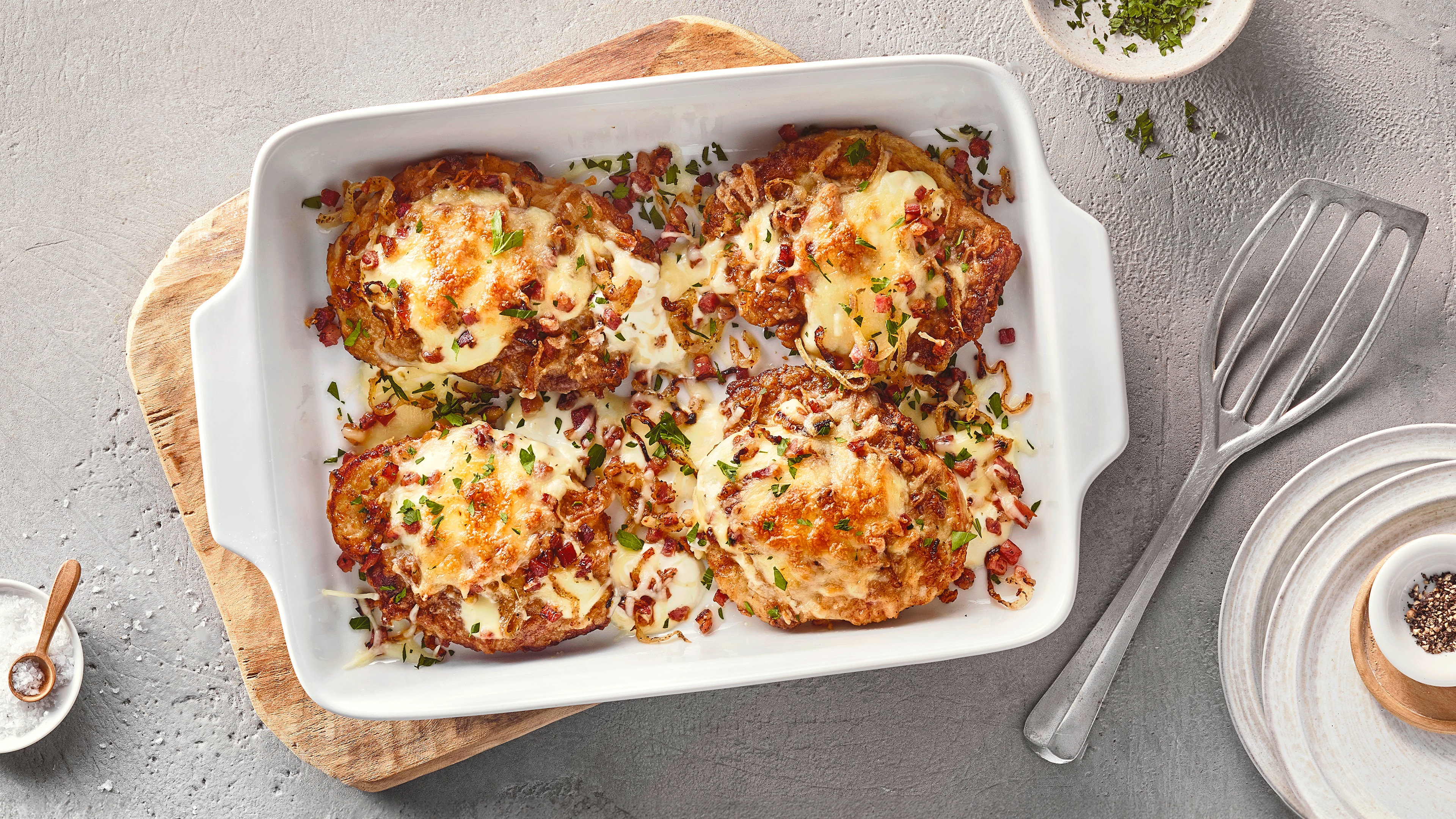 Eine Auflaufform mit vier gebräunten Fleischstücken, überbacken mit Käse, kleinen roten Würfeln & grünen Kräutern.