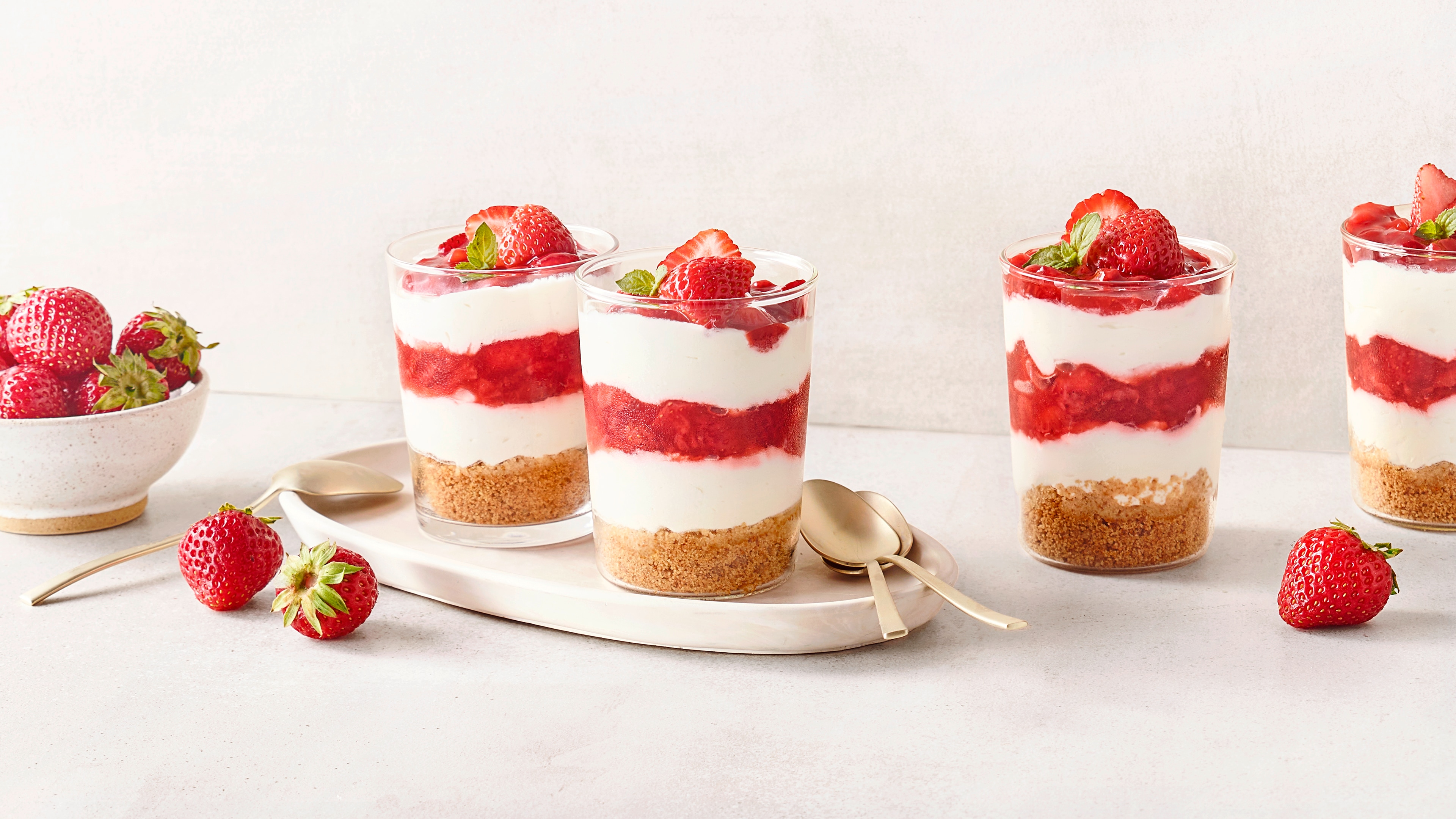 Dessertgläser mit Schichten aus Creme, Erdbeeren und Keksboden, garniert mit frischen Erdbeeren und Minze.