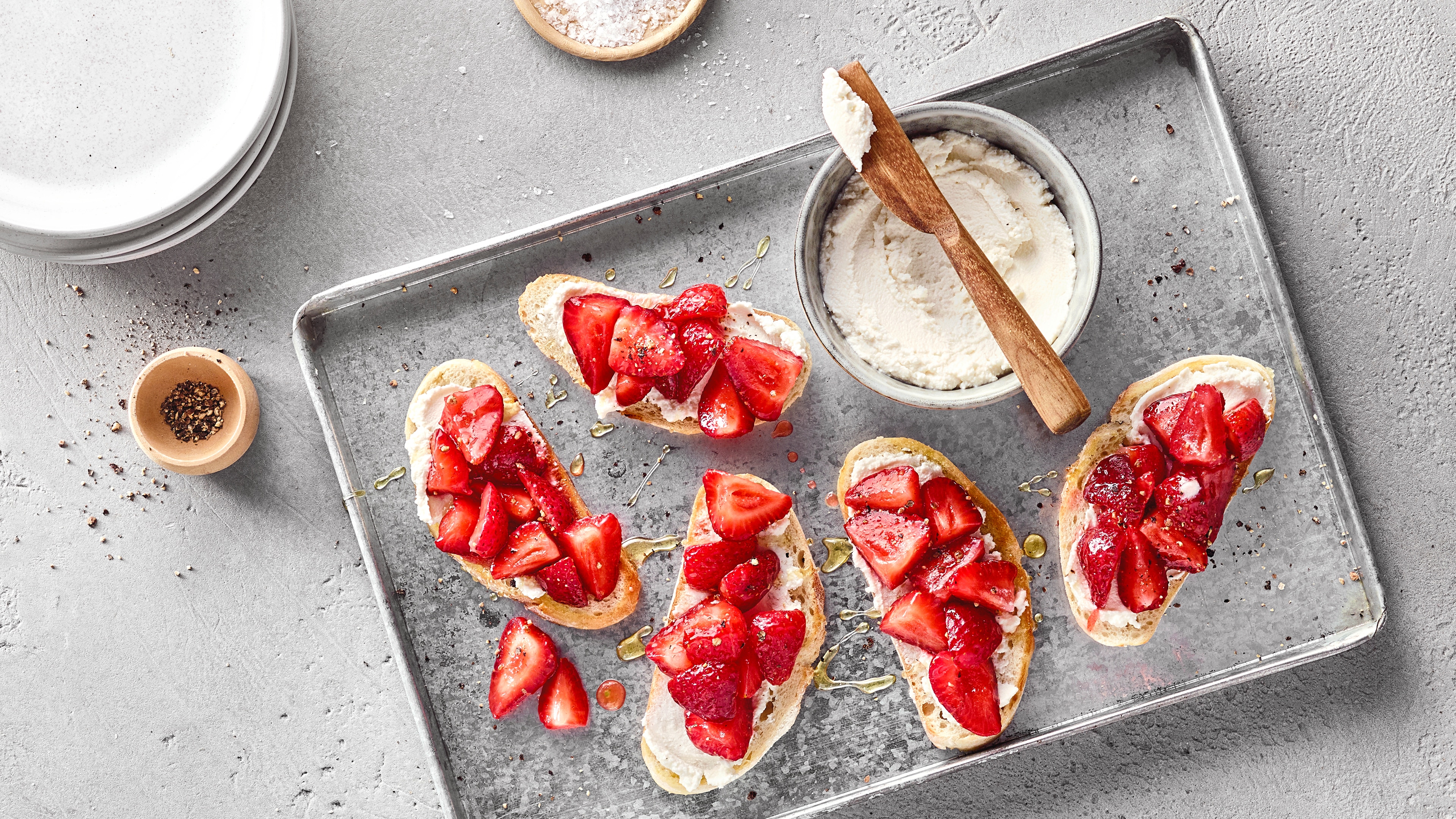 Vier Crostini mit Frischkäse und frischen Erdbeeren auf einem Backblech, garniert mit Honig; daneben eine Schale mit Frischkäse.