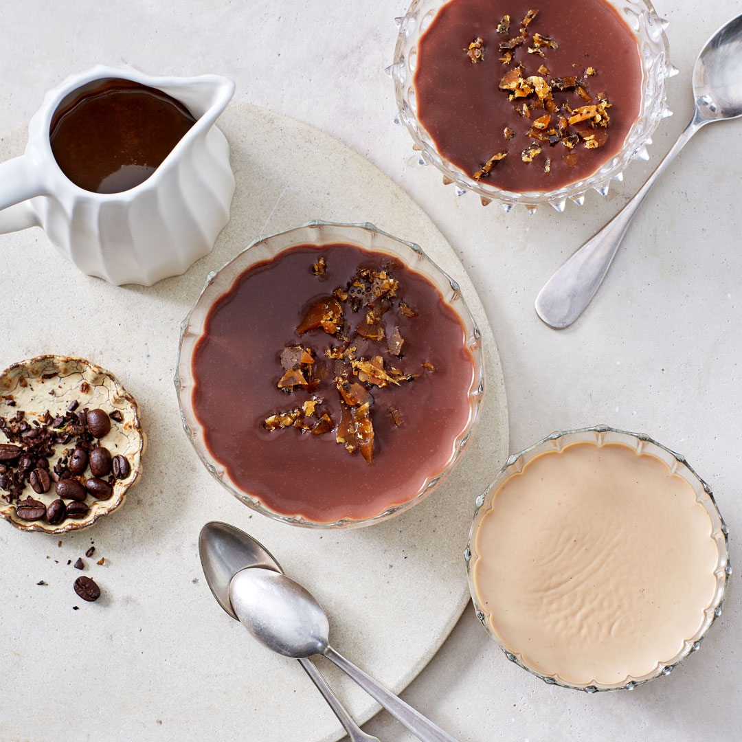 Glasschälchen, gefüllt mit heller, fester Creme,  getoppt mit einer dunklen Schokoschicht und Karamellstückchen