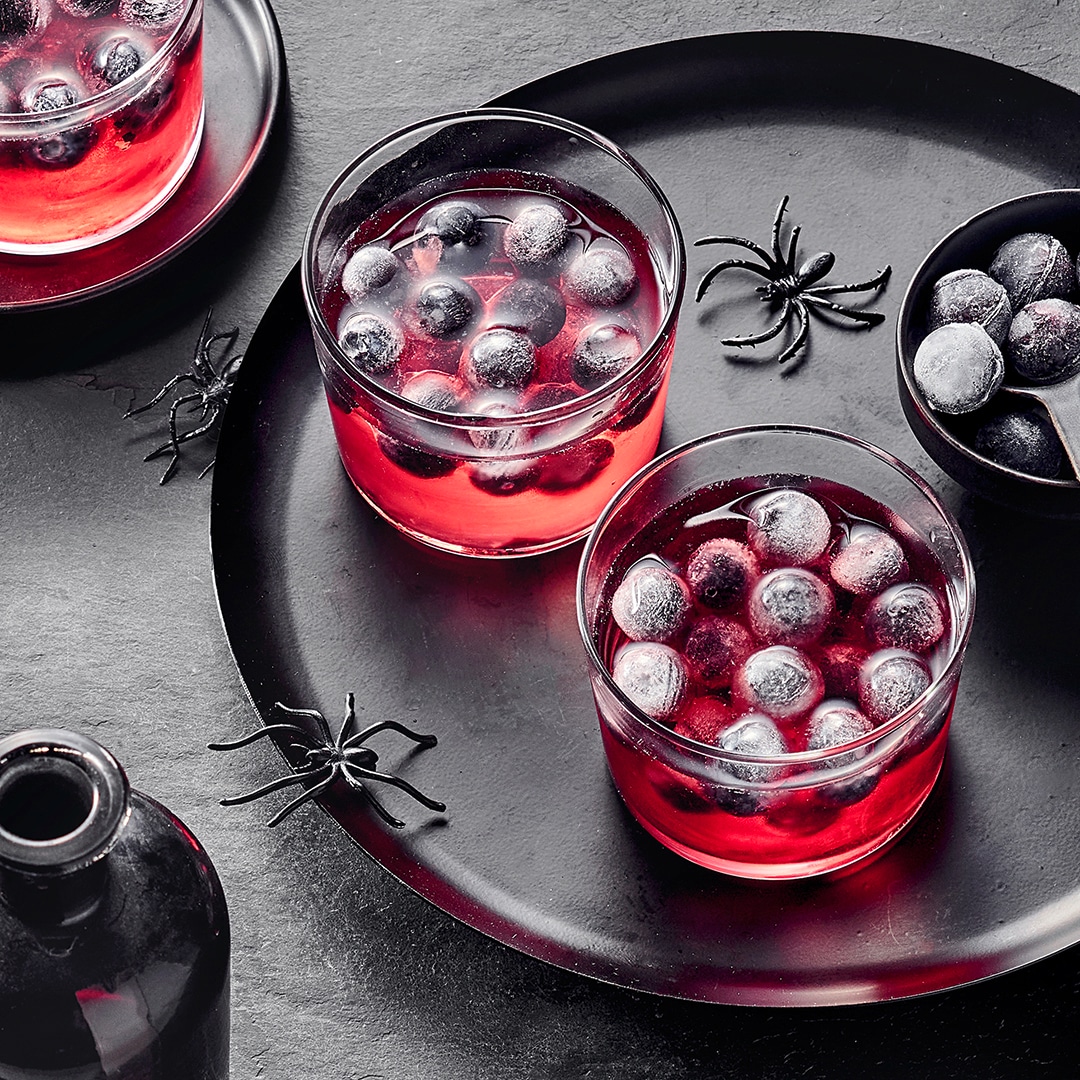 Gläser mit roter Flüssigkeit und gefrorenen Eiskugeln in Form von Augäpfeln, arrangiert auf einem Tablett mit dekorativen Spinnen.