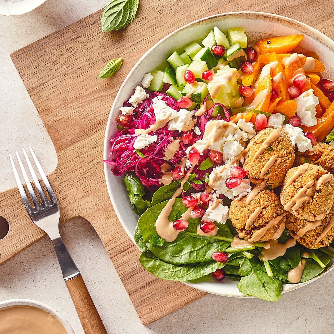 Eine Salat-Bowl mit Gurke, Paprika, Babyspinat und Rotkohl, getoppt mit Feta, Teigbällchen, Granatapfelkernen und heller Soße