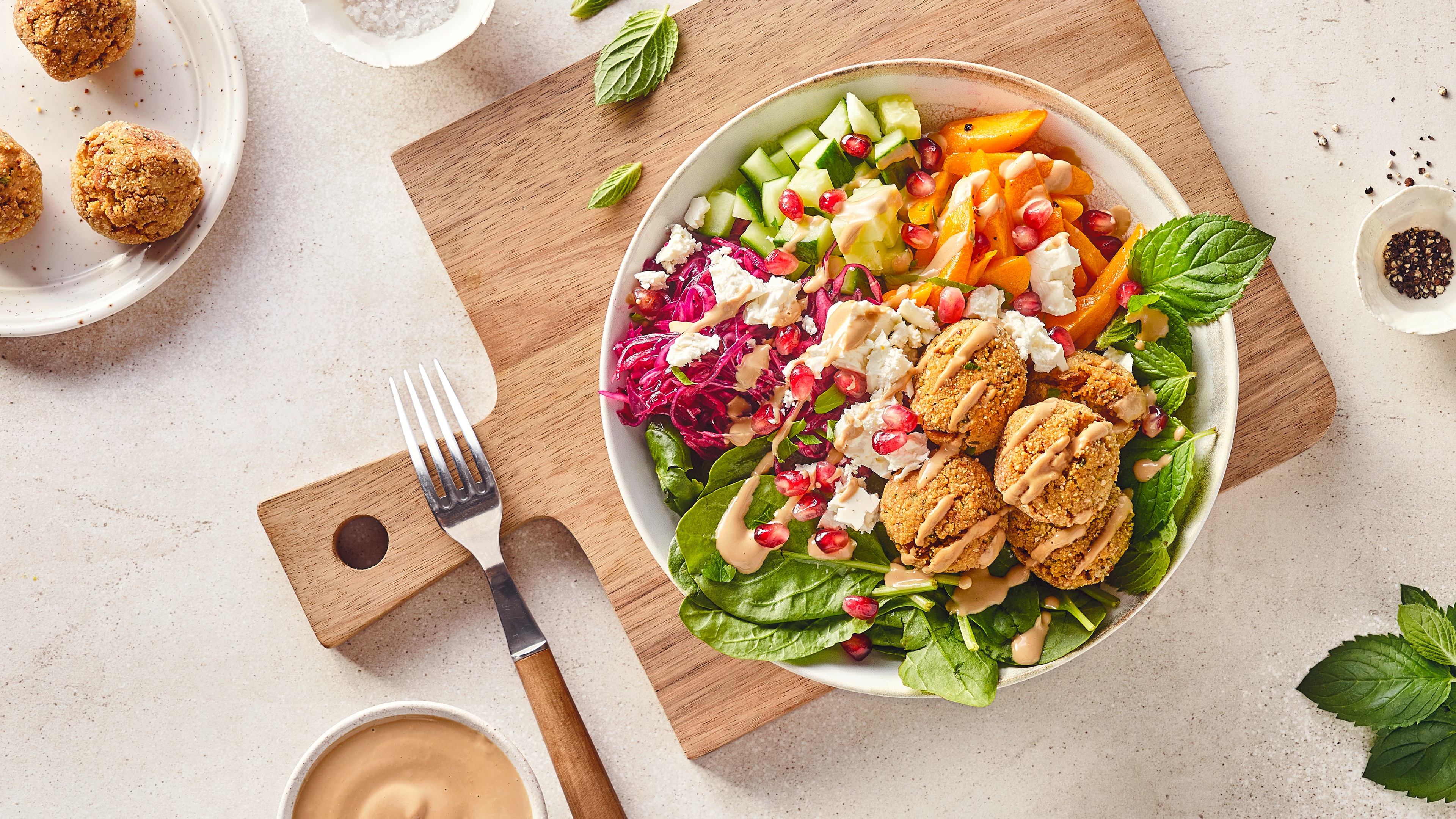 Eine Salat-Bowl mit Gurke, Paprika, Babyspinat und Rotkohl, getoppt mit Feta, Teigbällchen, Granatapfelkernen und heller Soße
