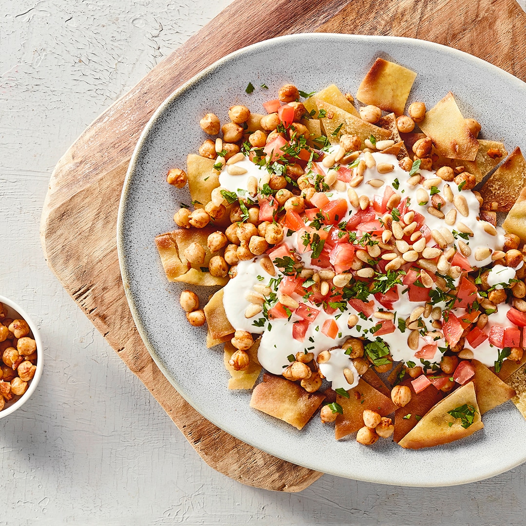 Ein Teller mit gebräunten Fladenbrotstücken, bedeckt mit weißen Cremeflecken, goldbraunen Kichererbsen, roten Würfeln und grünen Kräutern.