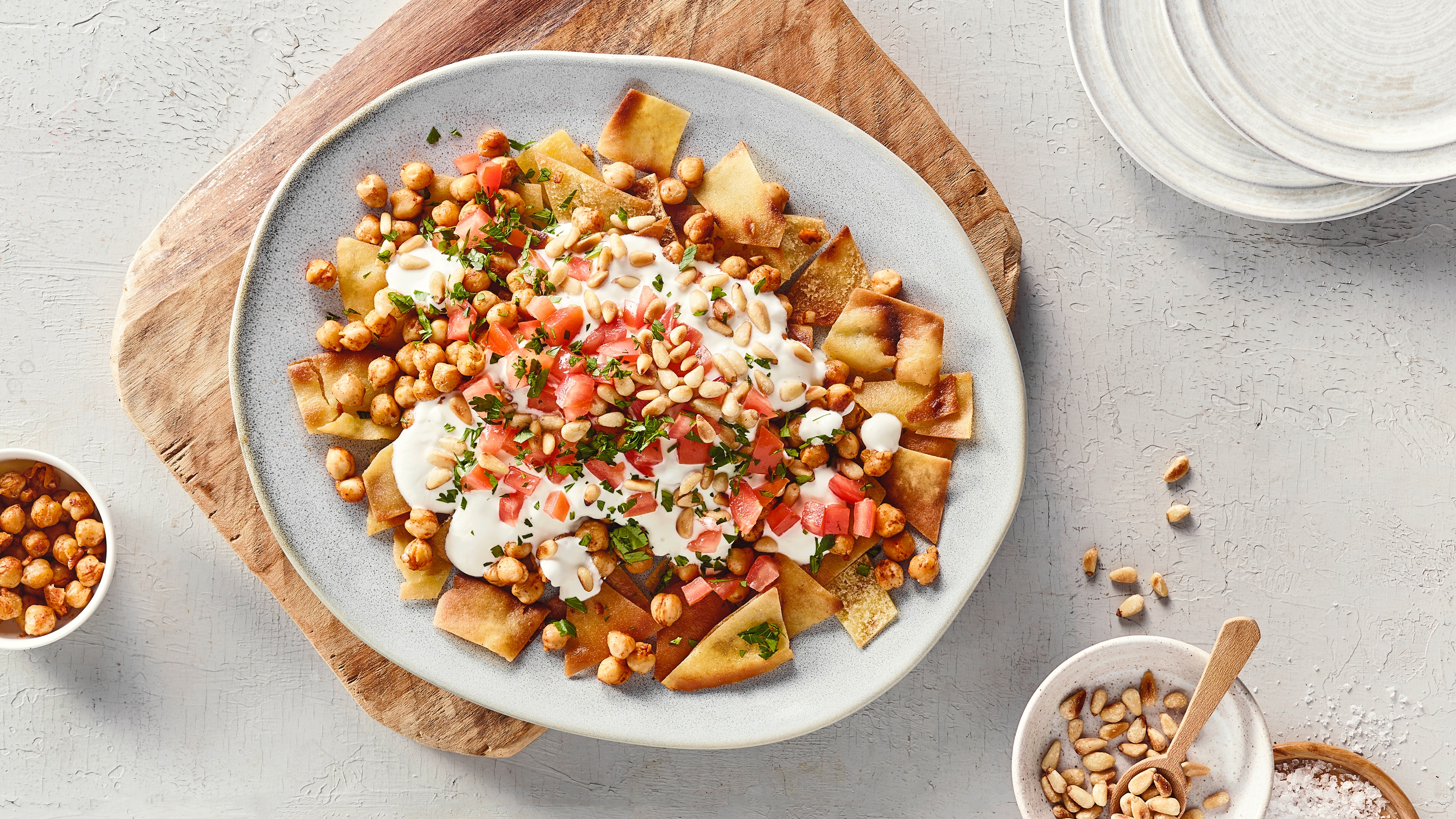Ein Teller mit gebräunten Fladenbrotstücken, bedeckt mit weißen Cremeflecken, goldbraunen Kichererbsen, roten Würfeln und grünen Kräutern.