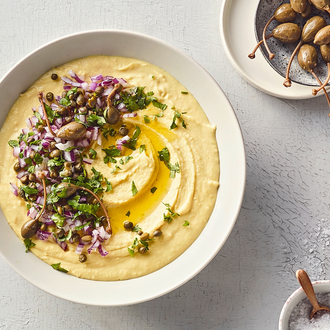 Eine Schale mit cremigem Aufstrich, garniert mit gehackten roten Zwiebeln, grünen Kräutern, Kapern und etwas Öl