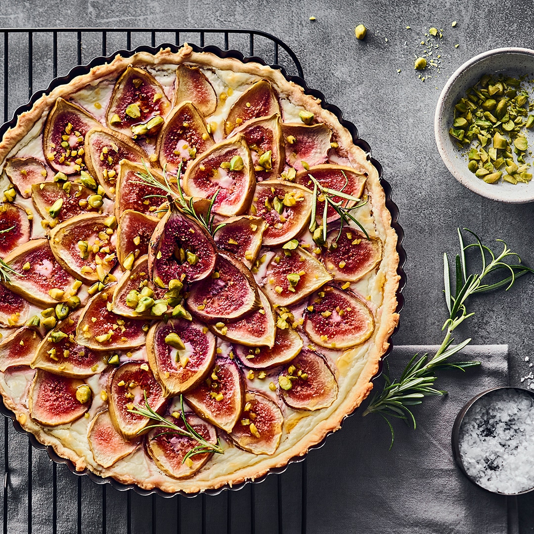 Eine runde Tarte mit Feigen und cremigem Ziegenfrischkäse auf einem Abkühlgitter, getoppt mit gehackten Pistazien.