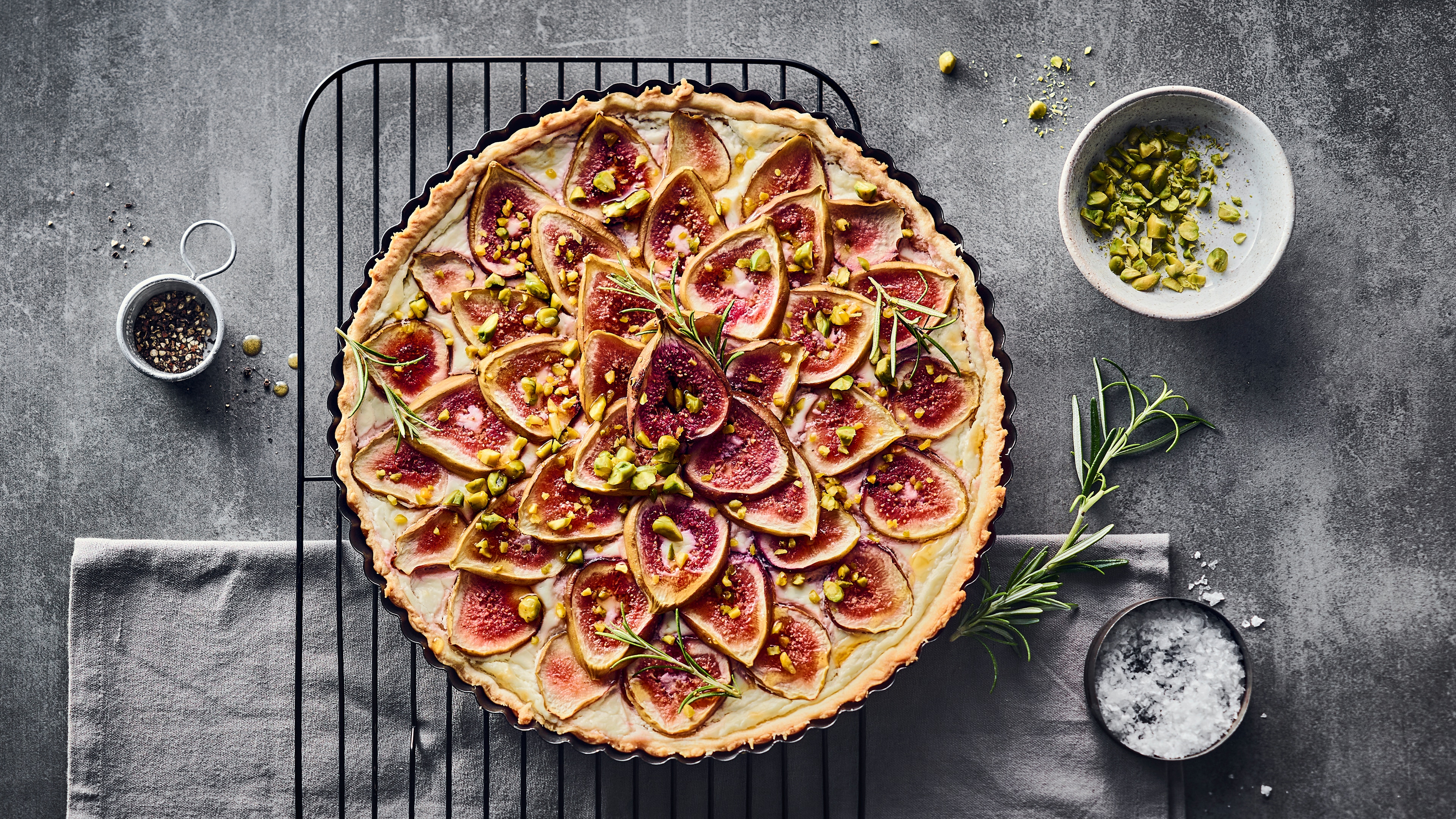 Rezept für eine Feigentarte mit Ziegenfrischkäse Eine runde Tarte mit Feigen und cremigem Ziegenfrischkäse auf einem Abkühlgitter, getoppt mit gehackten Pistazien.