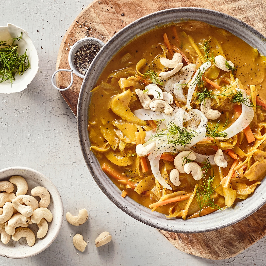 Eine Schüssel mit gelber Soße, darin orange Streifen, helle Scheiben, grüne Kräuter und ganze Cashewkerne