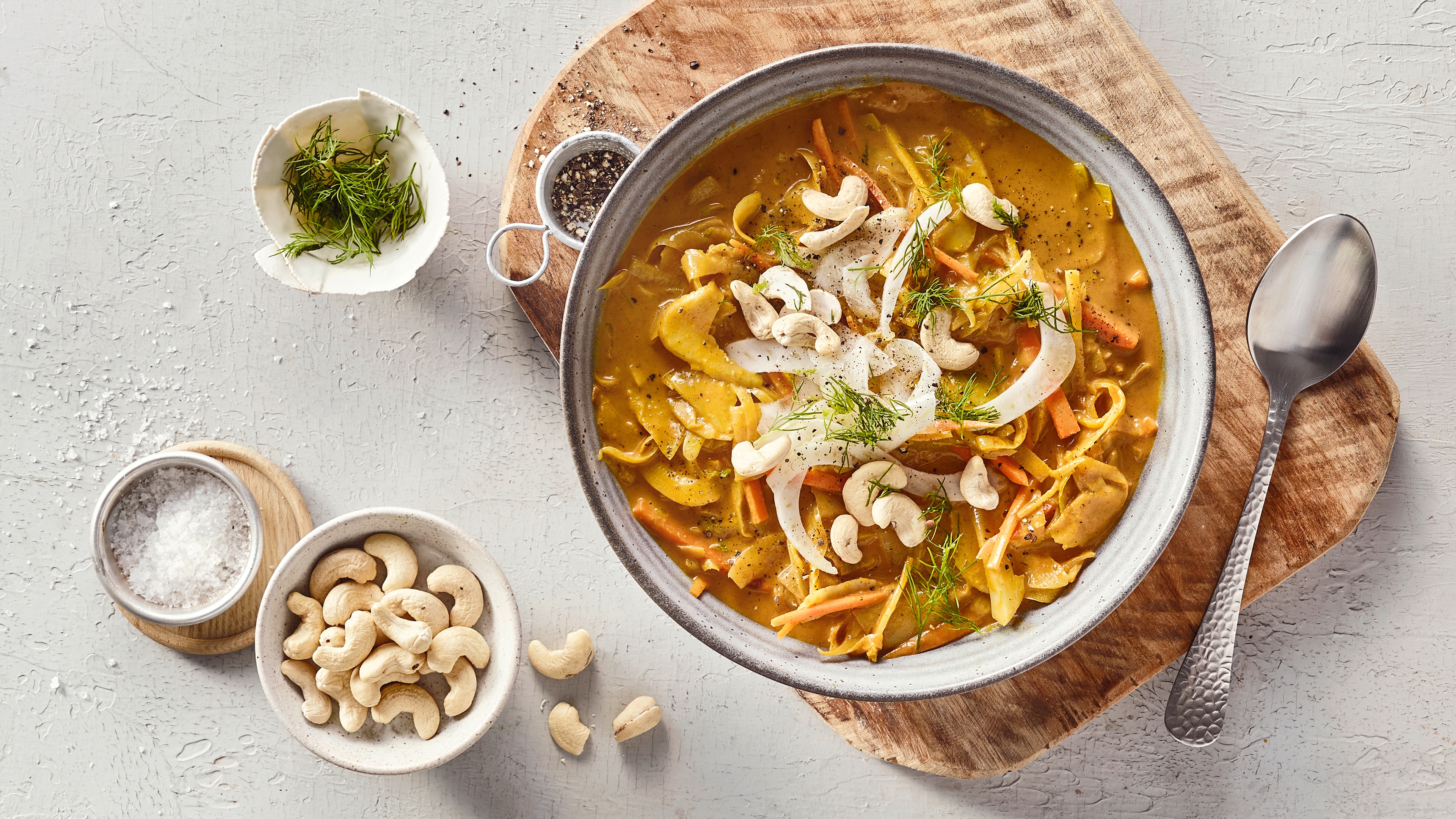 Eine Schüssel mit gelber Soße, darin orange Streifen, helle Scheiben, grüne Kräuter und ganze Cashewkerne