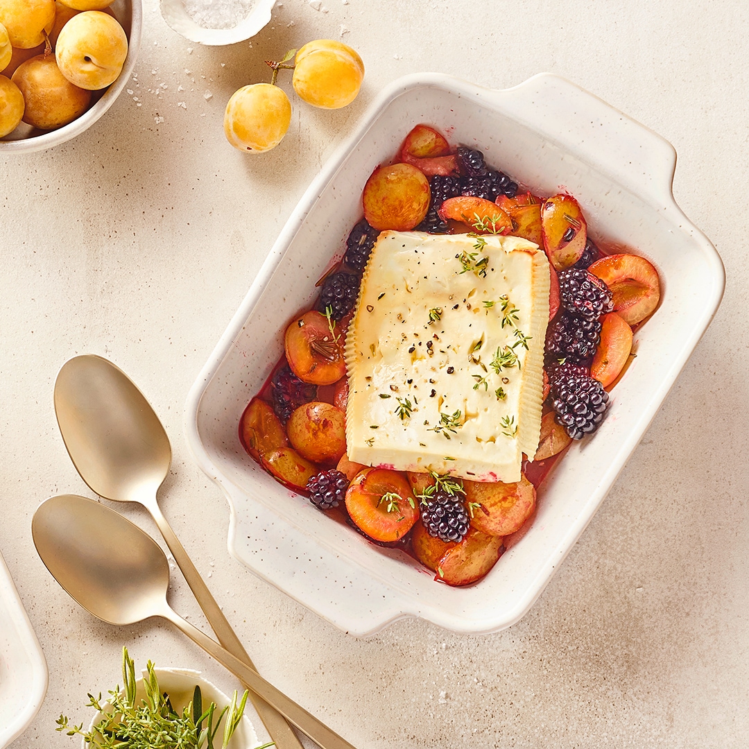 Auflaufform mit gebackenem Feta auf Mirabellen und Brombeeren, garniert mit Kräutern, daneben frische Mirabellen und Servierlöffel.