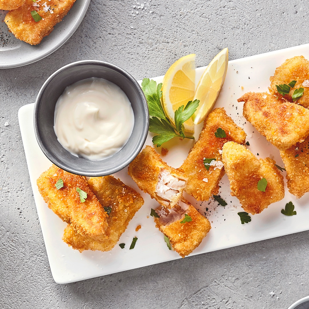 Ein Teller mit goldbraun frittierten Fischnuggets, daneben eine Schale mit hellem Dip.
