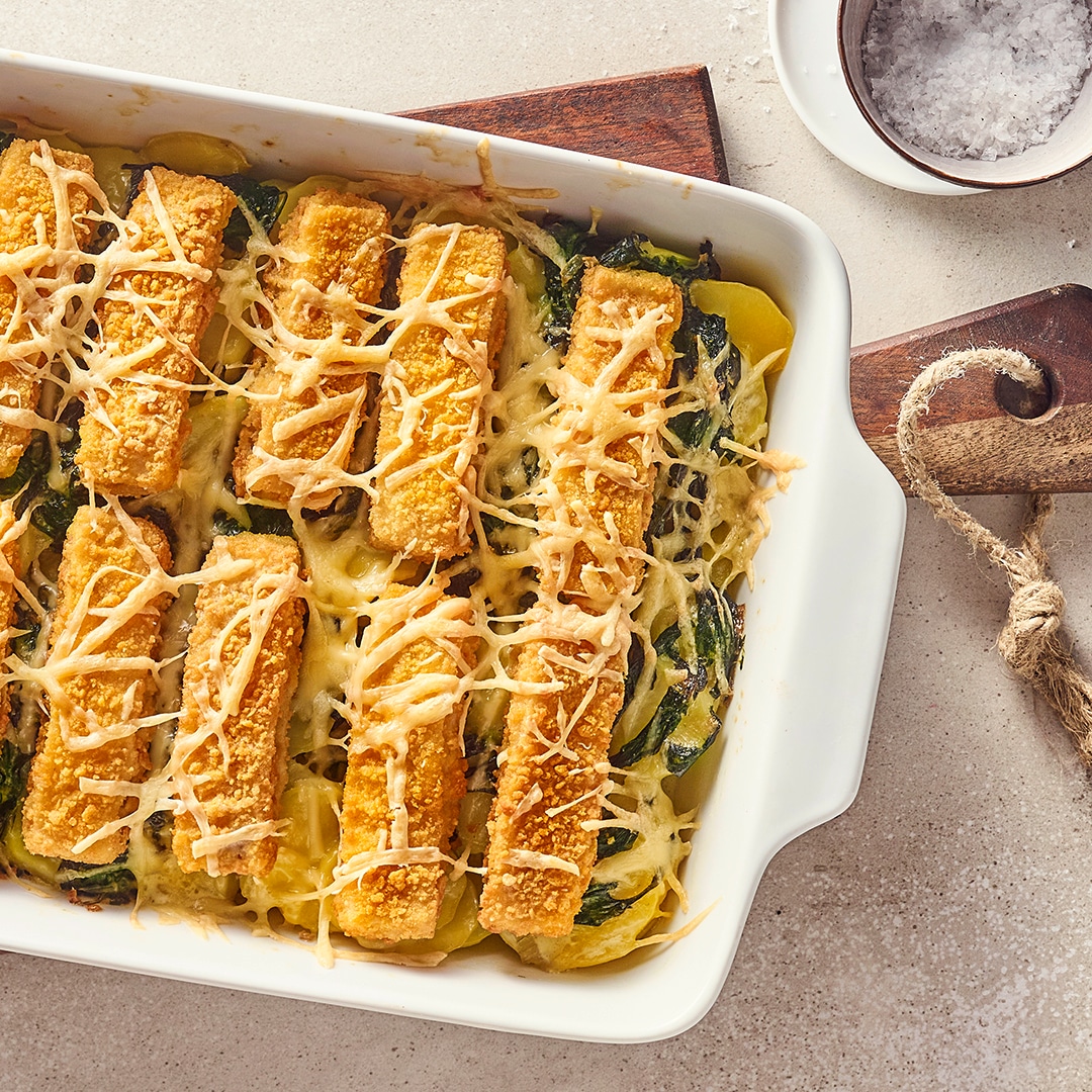 Eine Keramikauflaufform mit Kartoffelscheiben, Spinat und knusprigen Fischstäbchen, überbacken mit Käse