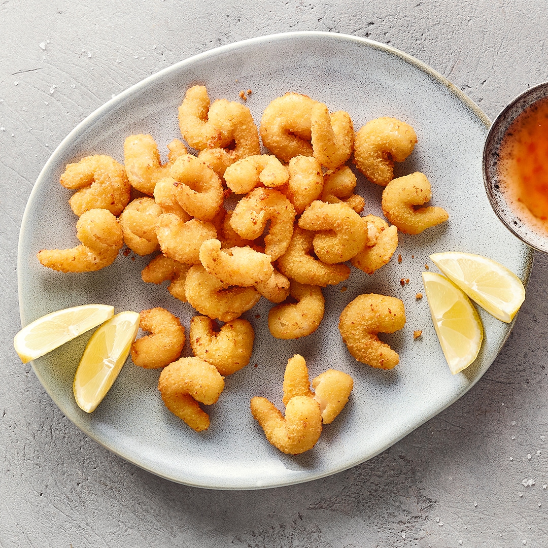 Ein Teller mit panierten Garnelen, umgeben von Zitronenspalten und einer Schale mit orangefarbener Soße am Rand.