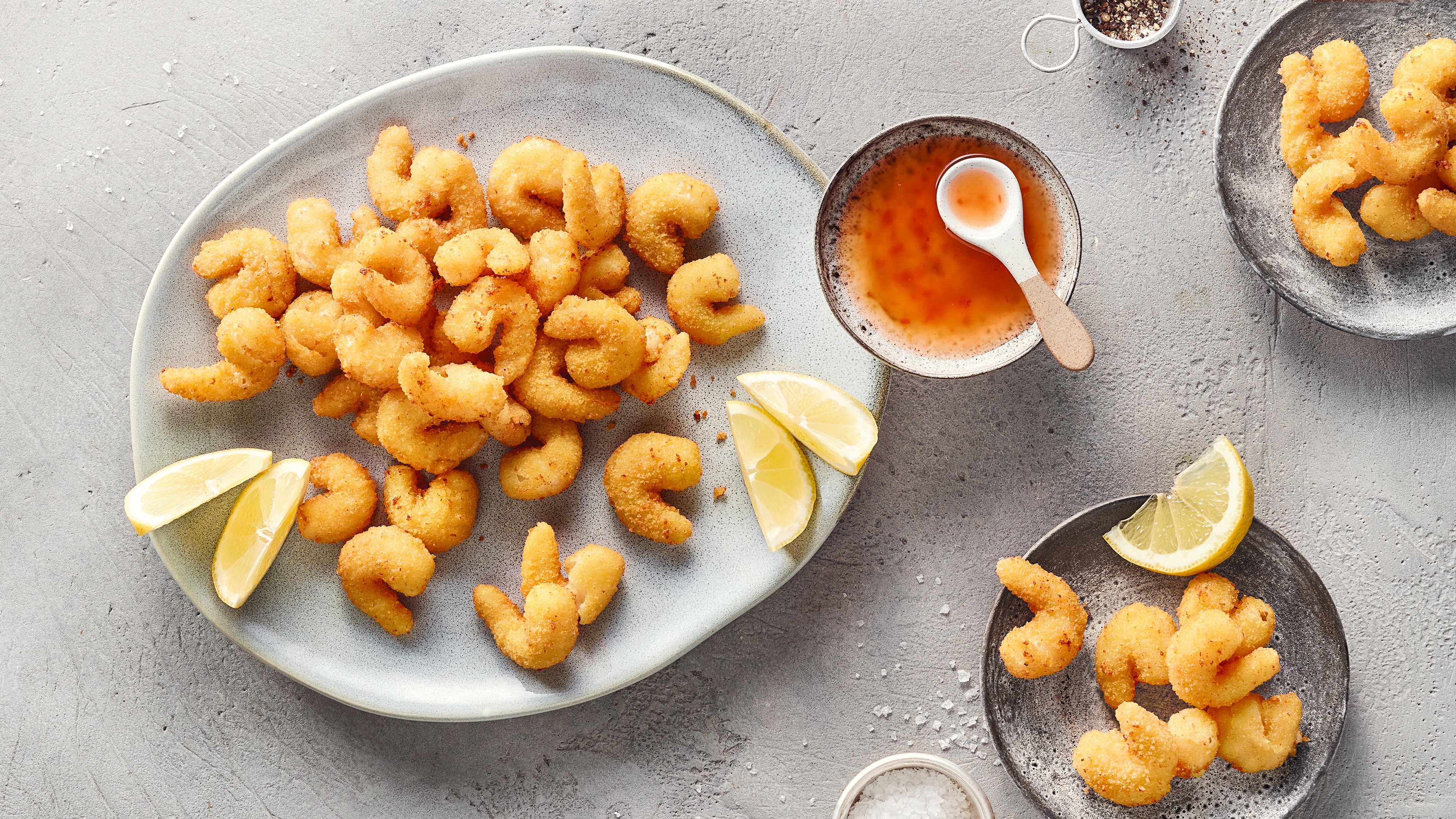Ein Teller mit panierten Garnelen, umgeben von Zitronenspalten und einer Schale mit orangefarbener Soße am Rand.
