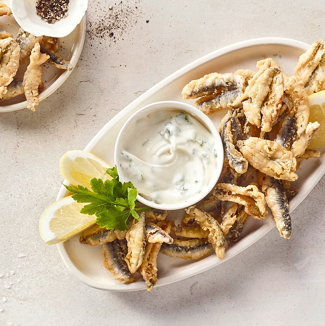 Länglicher Teller mit kleinen panierten Fischen, daneben ein heller Dip mit Kräutern