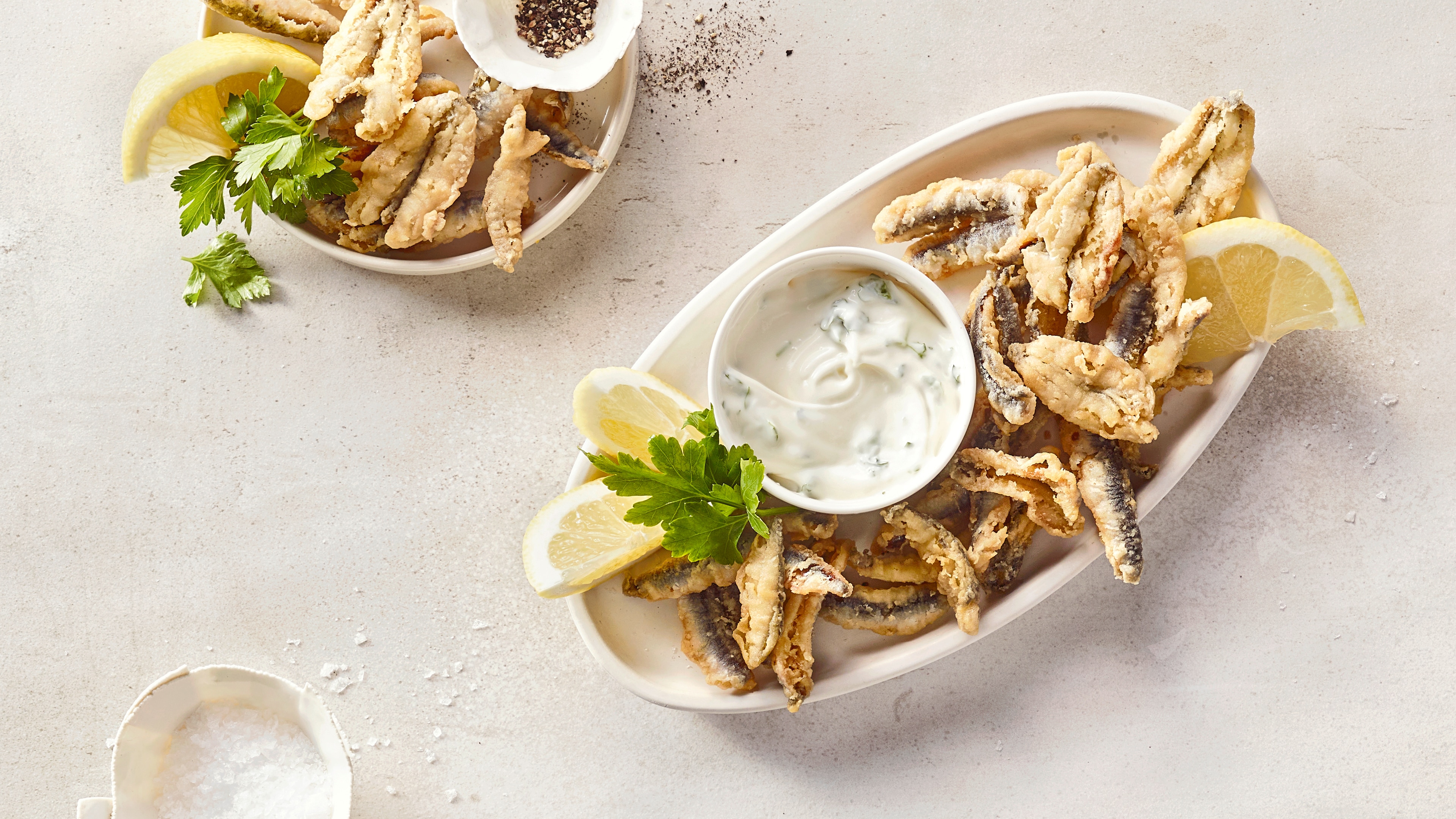 Länglicher Teller mit kleinen panierten Fischen, daneben ein heller Dip mit Kräutern