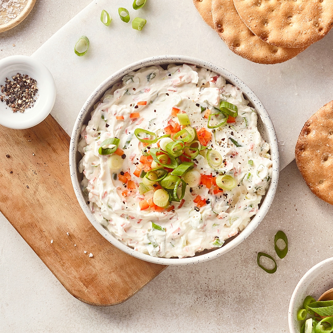 Eine Schale mit weißem Dip, darin grüne Frühlingszwiebelringe und kleine rote Stücke, bestreut mit schwarzem Pfeffer.