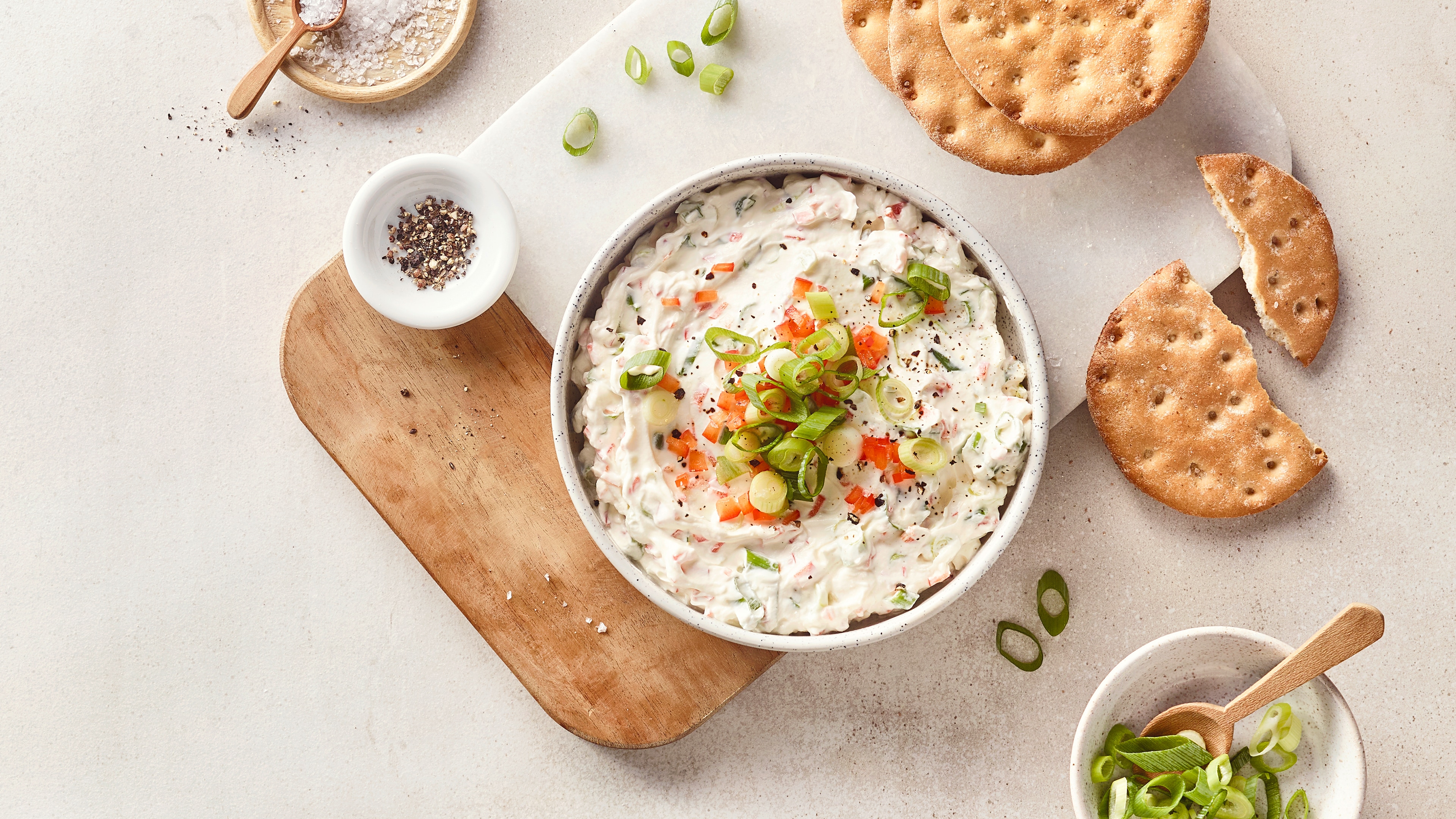 Eine Schale mit weißem Dip, darin grüne Frühlingszwiebelringe und kleine rote Stücke, bestreut mit schwarzem Pfeffer.