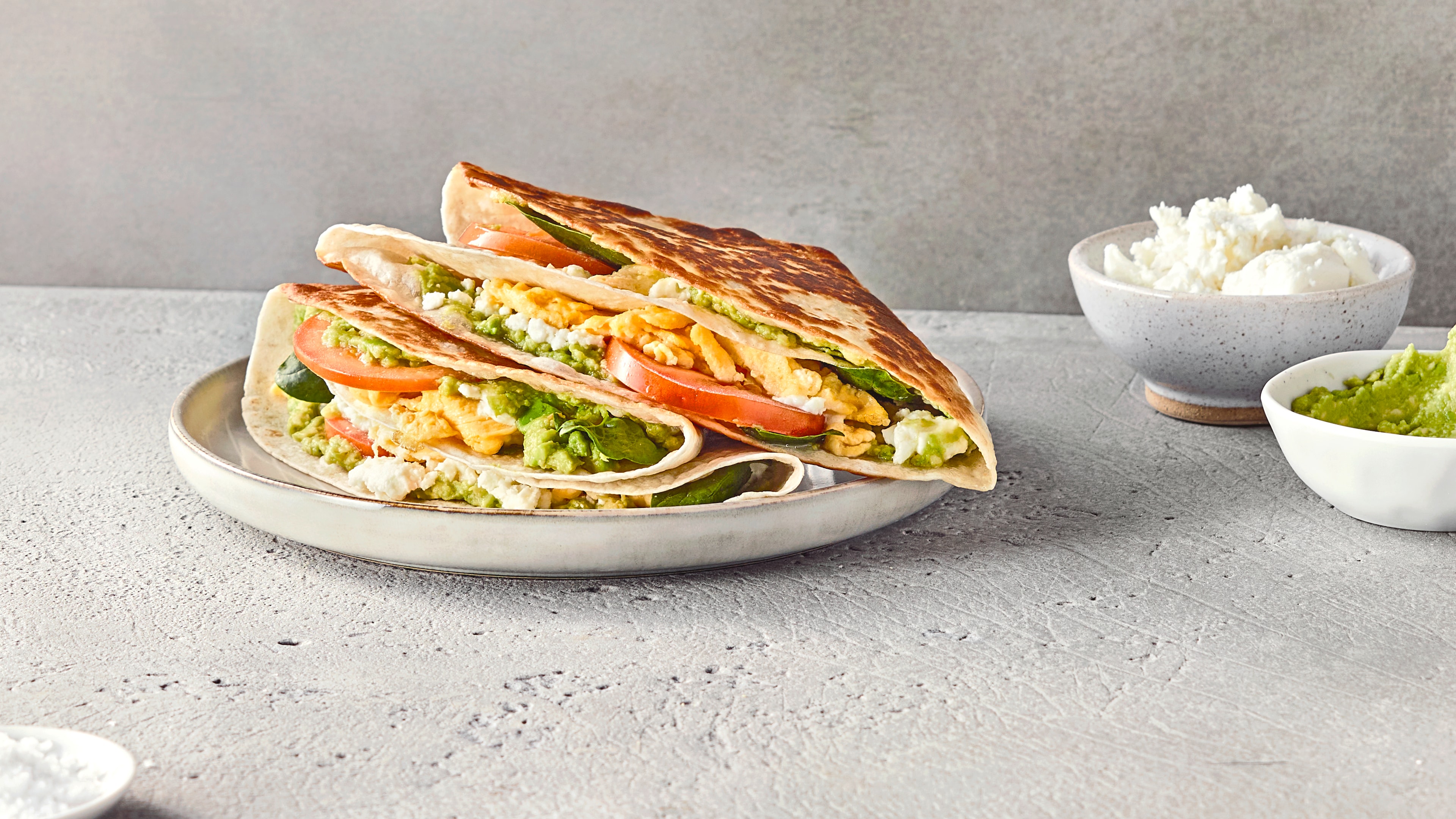 Rezept für Frühstückswrap Ein Teller mit braun angebratenen, gefalteten Wraps, gefüllt mit grüner Soße, Mozzarella, Salat, Tomate und Rührei