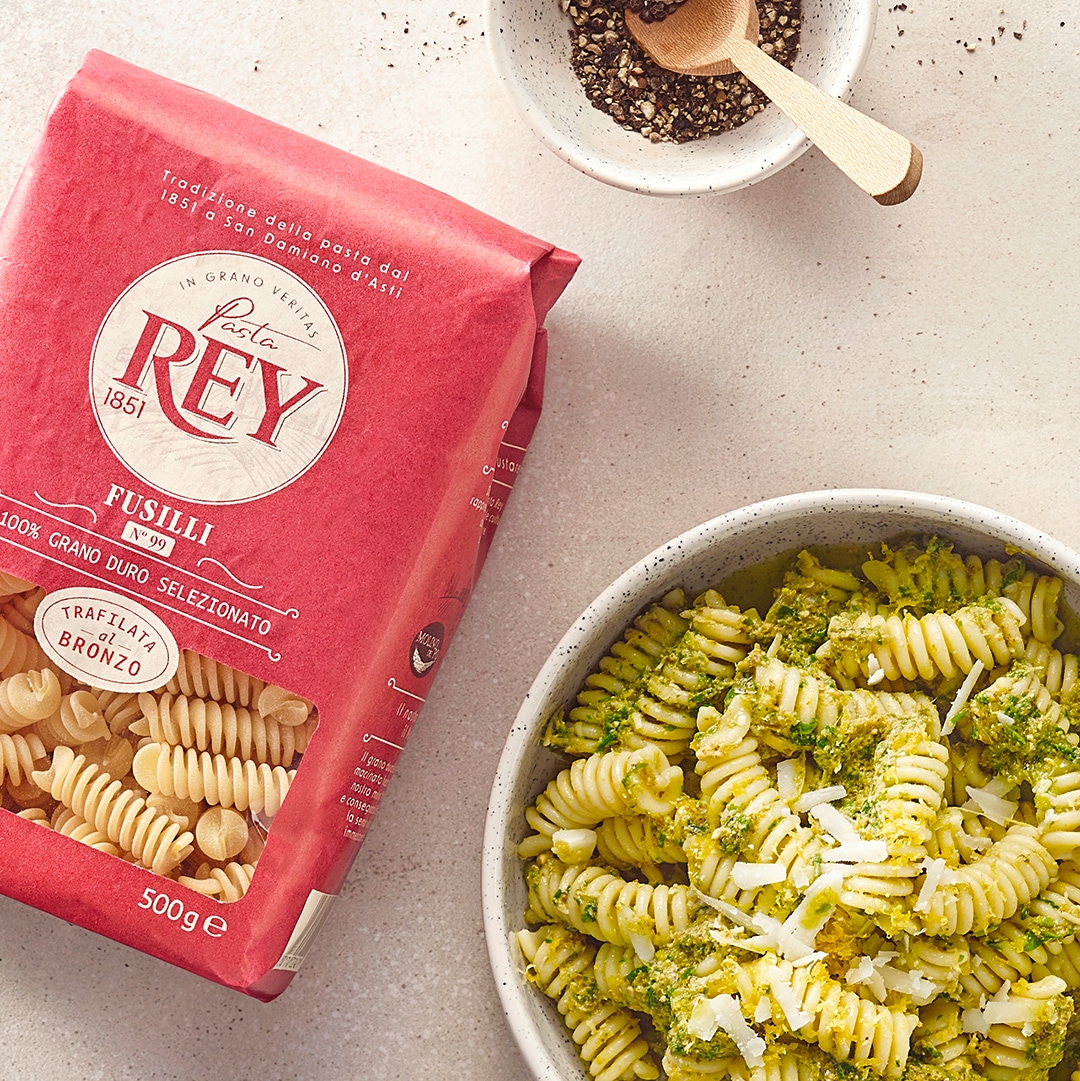 Schale mit gedrehten Nudeln, grüner Soße und geriebenem Käse. Daneben eine Packung Pasta Rey Fusilli.